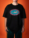 A person wearing an oversized black graphic T-shirt with a circular 'Bad Choices Good Stories' print in pink, teal, and white, paired with dark green athletic pants with yellow stripes, set against a solid orange background.