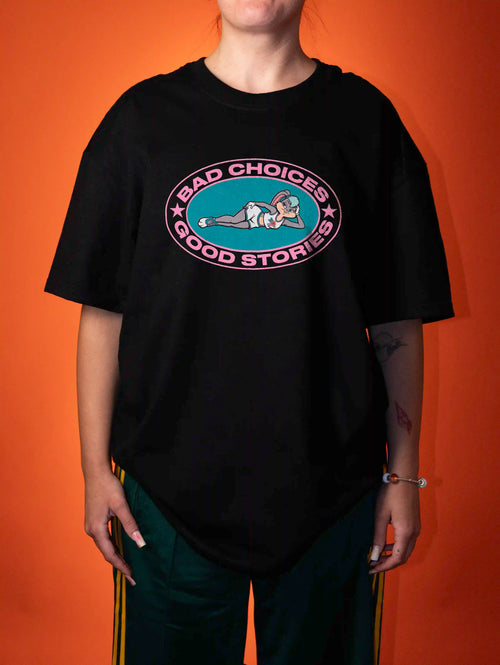A person wearing an oversized black graphic T-shirt with a circular 'Bad Choices Good Stories' print in pink, teal, and white, paired with dark green athletic pants with yellow stripes, set against a solid orange background.