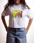 A person wearing a white cropped T-shirt with a large colorful MTV logo graphic on the front, paired with light blue jeans, against a plain light background.