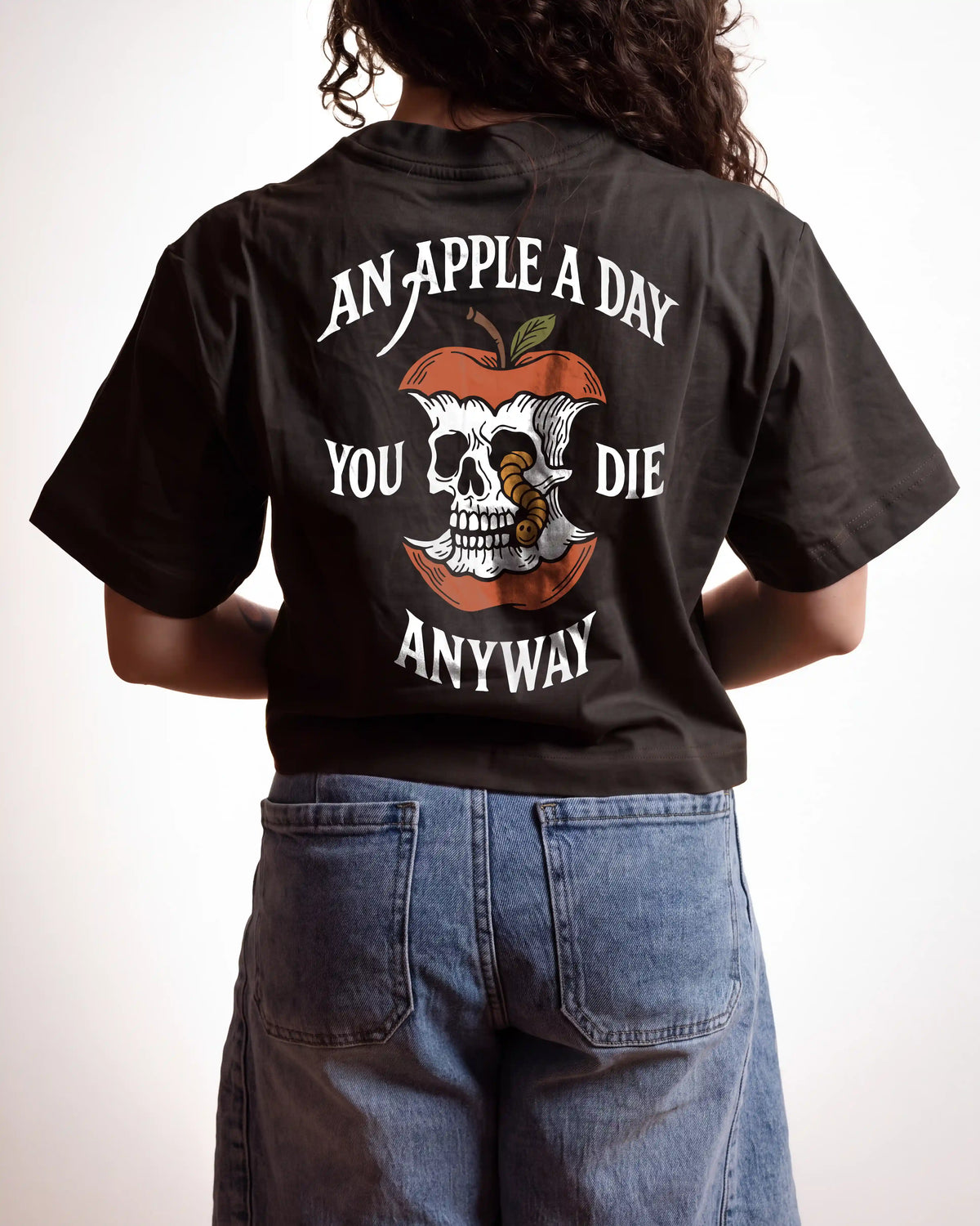 A person shown from the back wearing a black graphic T-shirt and blue jeans against a plain light background. The shirt features a large apple-and-skull design with the text 'AN APPLE A DAY YOU DIE ANYWAY'.