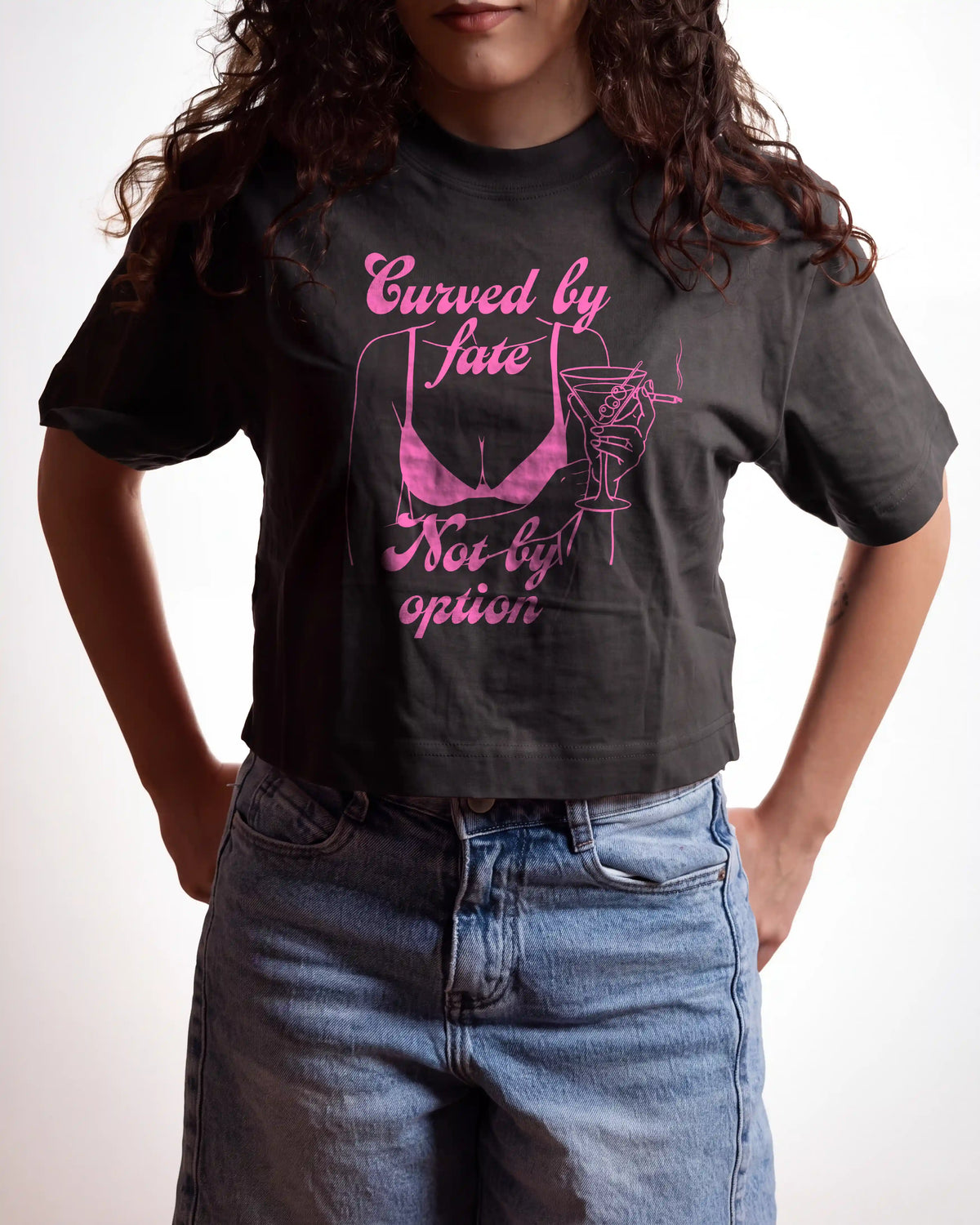 A woman wearing a black graphic t-shirt with pink text and a bikini/cocktail illustration, paired with light blue jeans, against a plain white background.