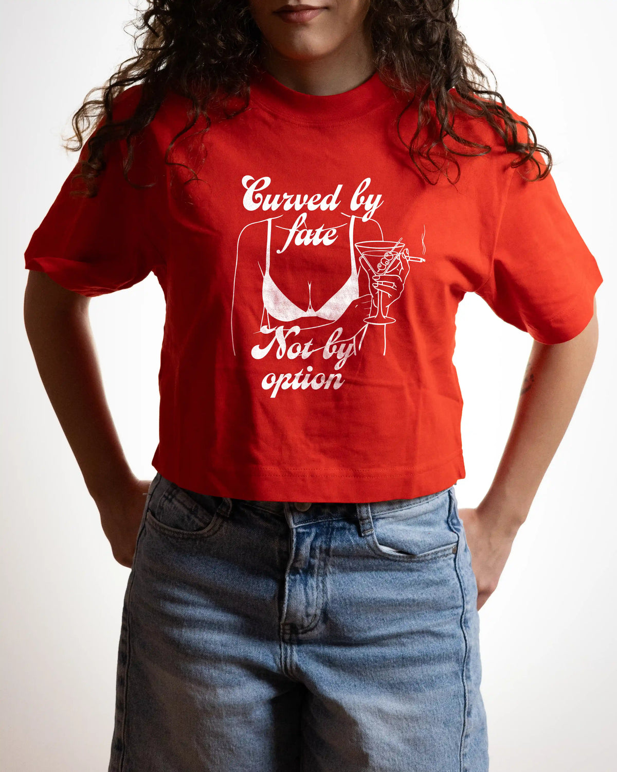 A person wearing a bright red graphic T-shirt with white text reading 'Curved by fate Not by option' and a white line drawing of a bra, paired with blue jeans against a plain light background.