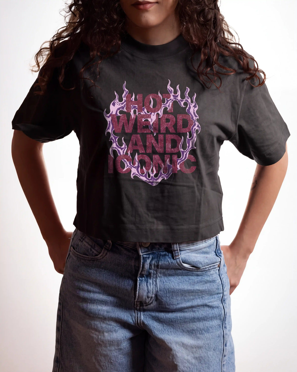 A person wearing a black graphic T-shirt with a pink and purple flame-style heart design and the words 'HOT WEIRD AND ICONIC,' paired with light blue jeans against a plain white background.