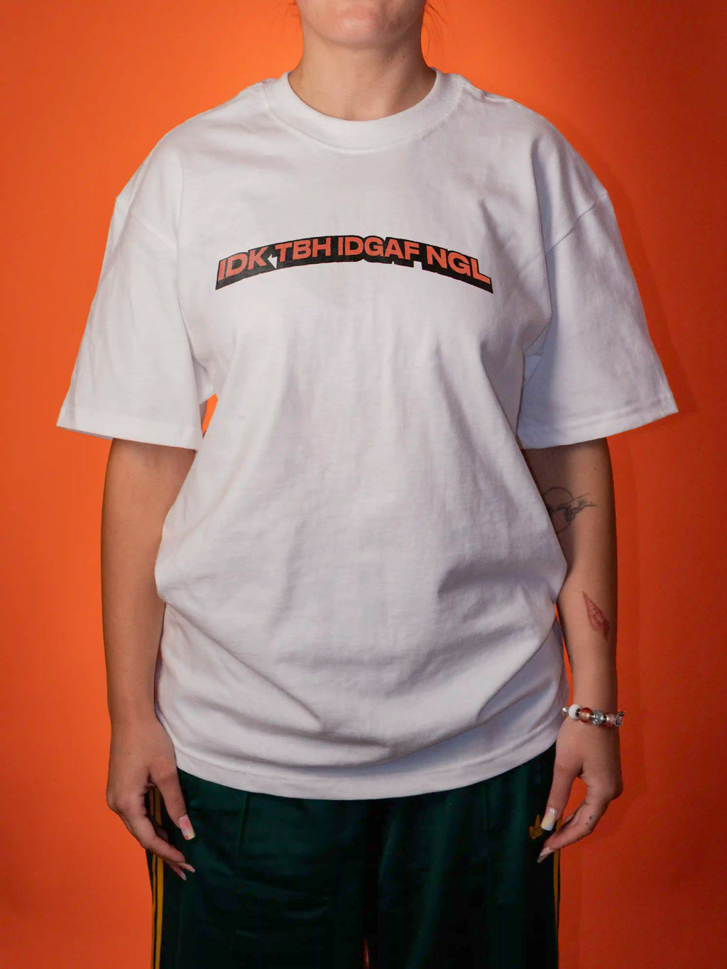 A person wearing an oversized white graphic T-shirt with the text 'IDK TGH IDGAF NGL' printed across the chest in black and orange, posed against a solid orange background.