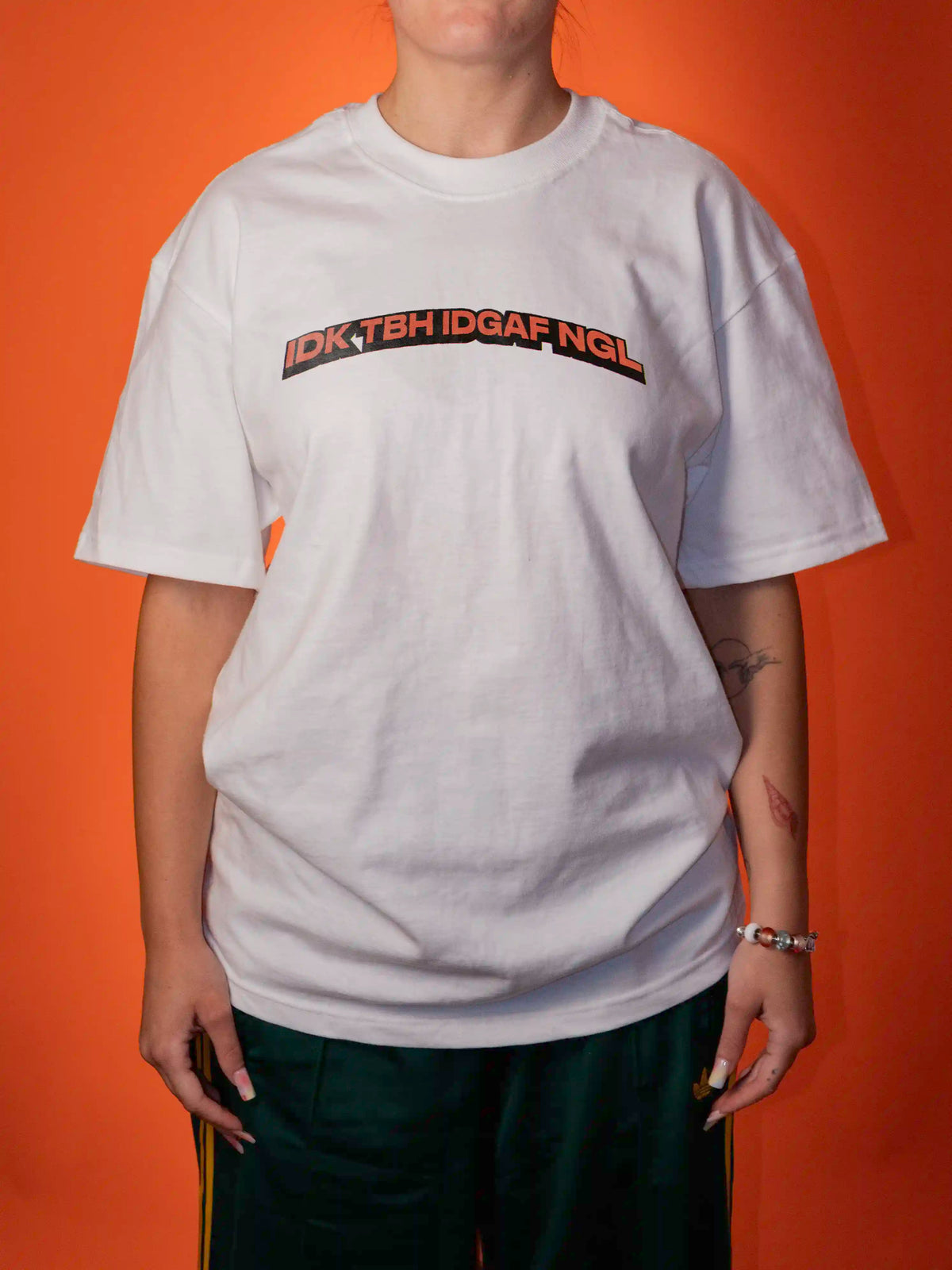 A person wearing an oversized white graphic T-shirt with the text 'IDK TGH IDGAF NGL' printed across the chest in black and orange, posed against a solid orange background.