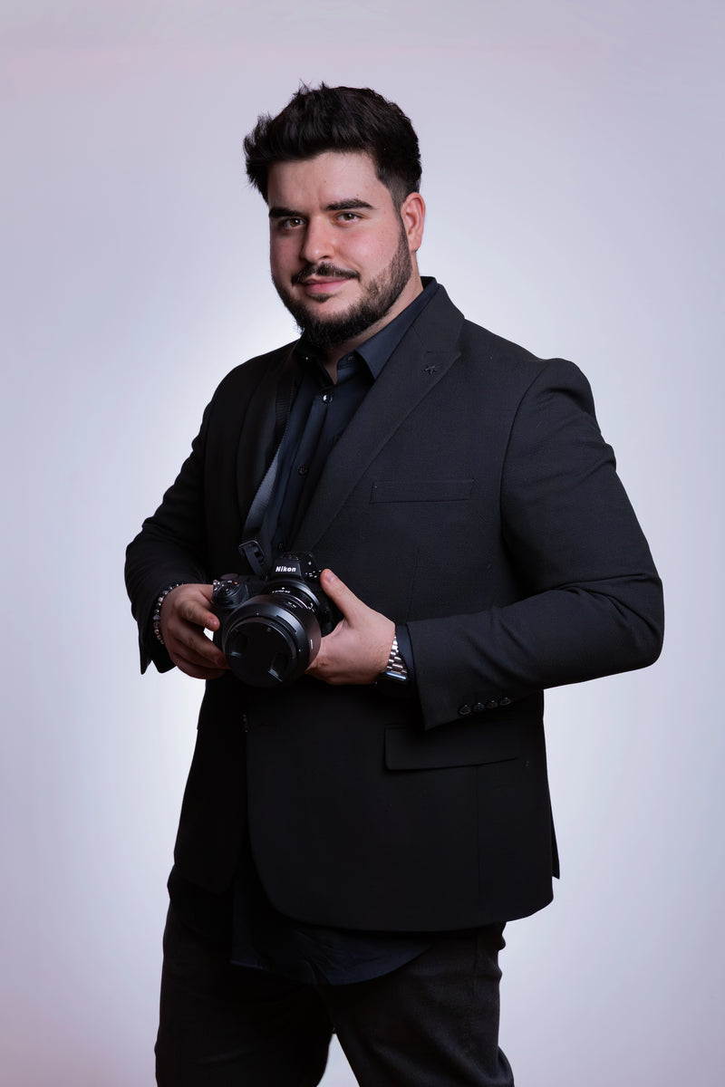 Man in a black suit holding a camera against a light gray background