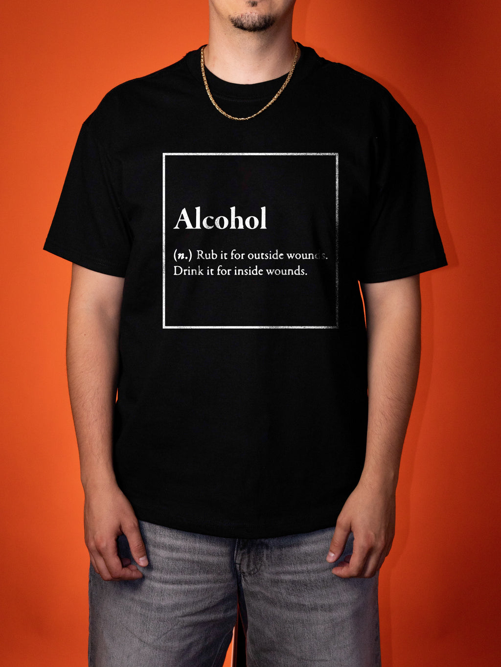 A man wearing a black graphic t-shirt with a white printed definition of the word 'Alcohol' on the chest, posed against a solid orange background.