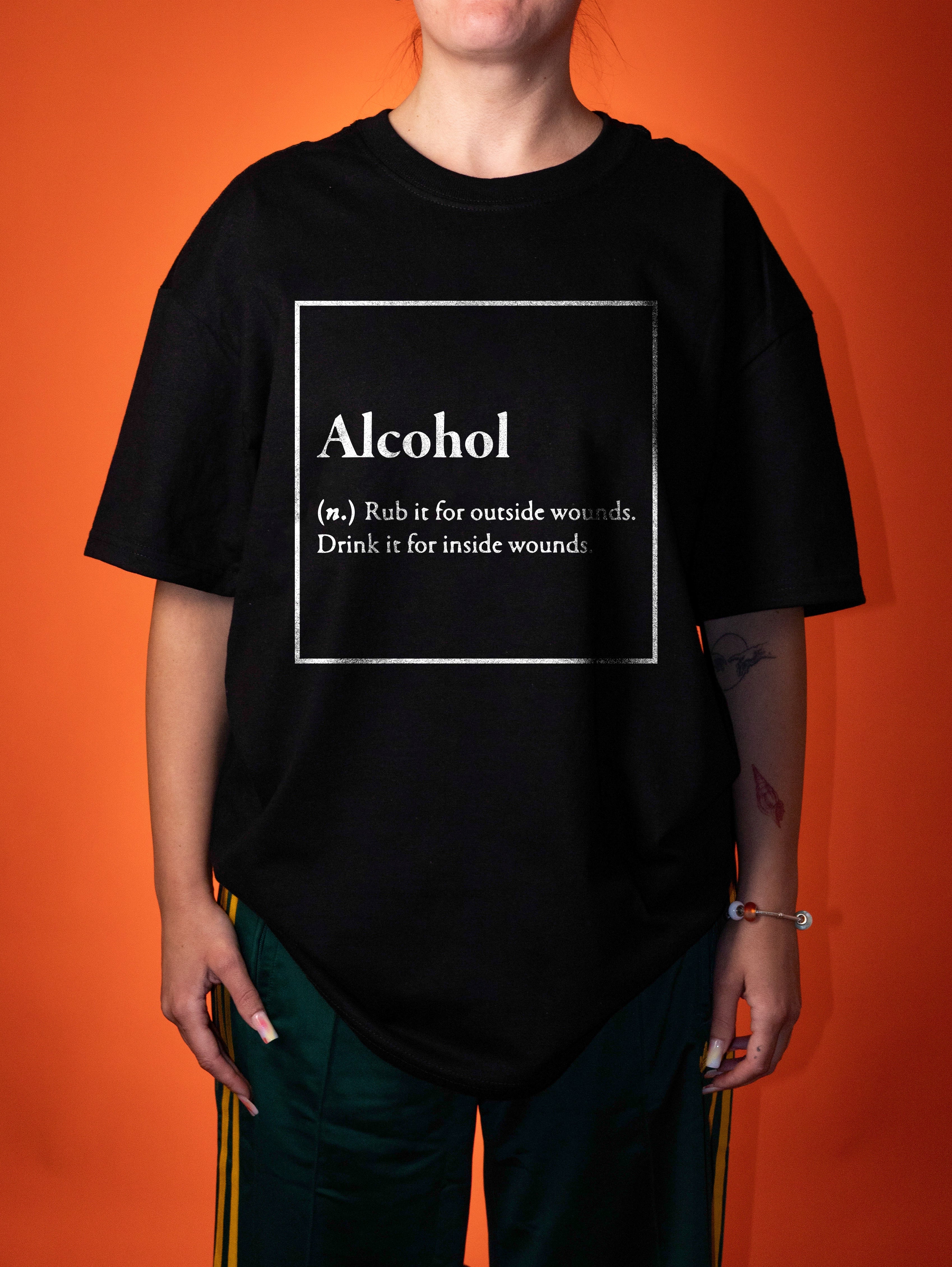 A person wearing an oversized black graphic T-shirt with a white printed definition of the word 'Alcohol' on the front, posed against a solid orange background.