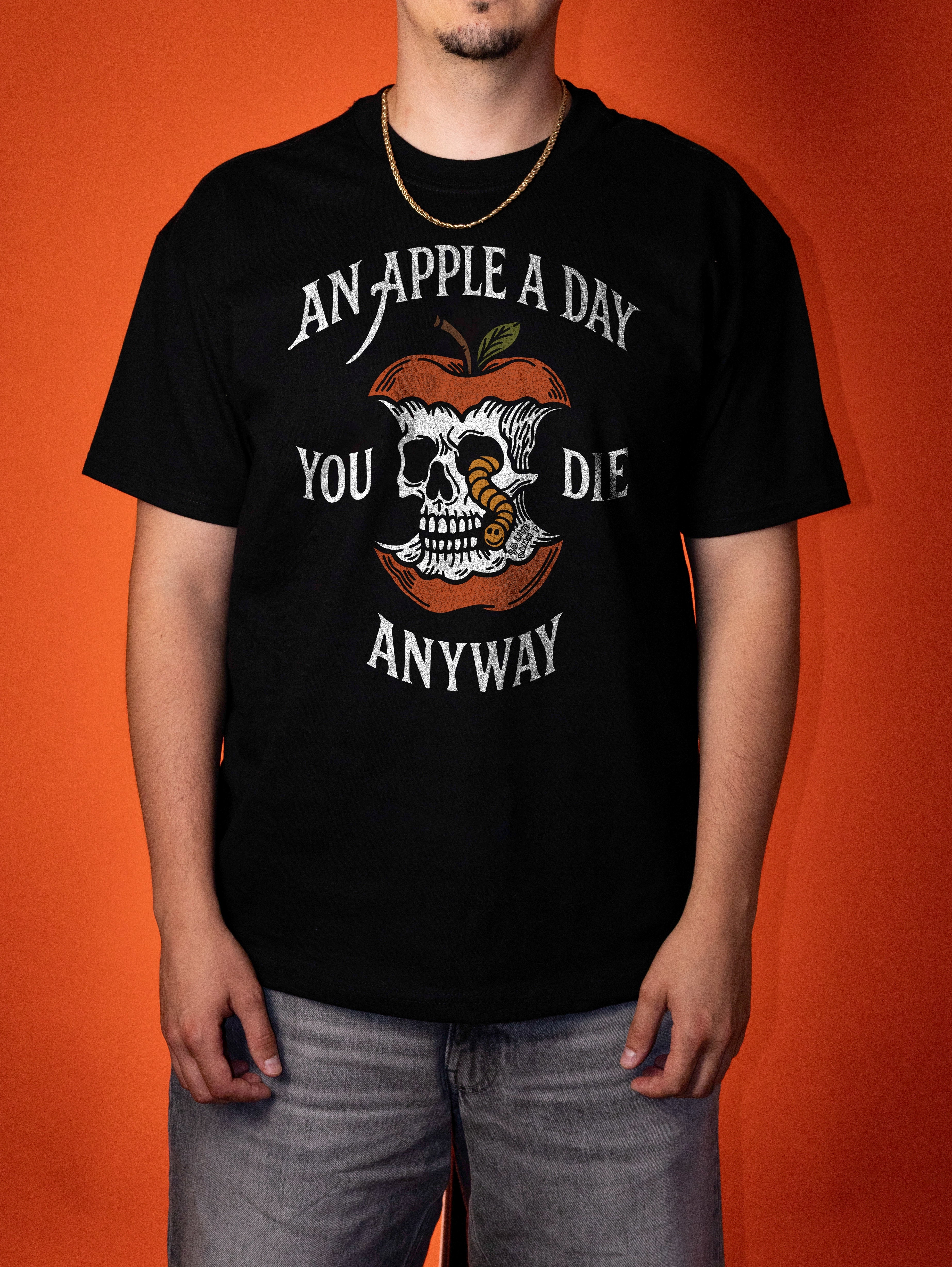 A man wearing a black graphic t-shirt with a skull and apple design and the text 'AN APPLE A DAY YOU DIE ANYWAY', posed against a solid orange background.