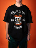 A person wearing an oversized black graphic t-shirt with a skull and apple design and the text 'AN APPLE A DAY YOU DIE ANYWAY', posed against a solid orange background.