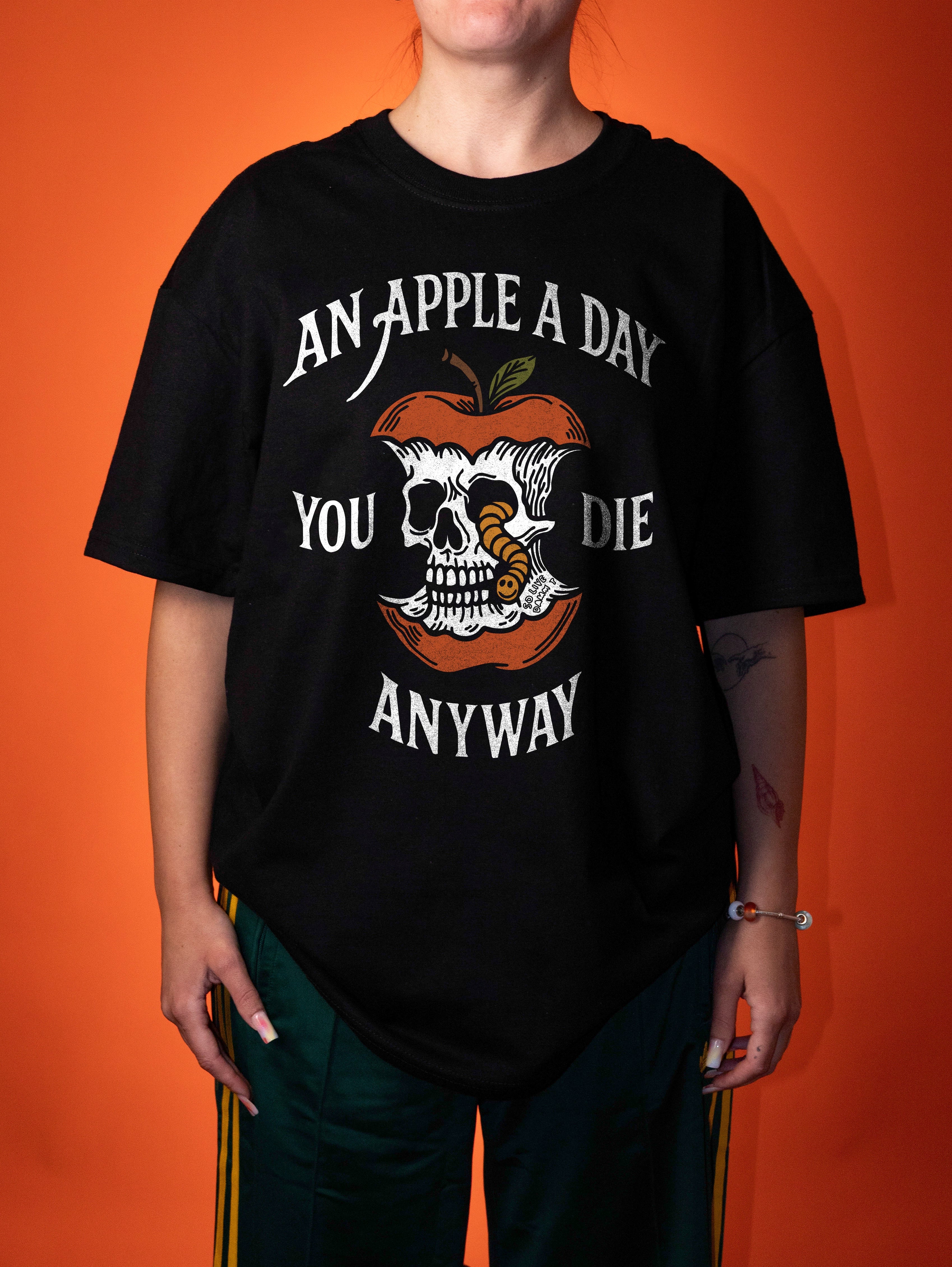 A person wearing an oversized black graphic t-shirt with a skull and apple design and the text 'AN APPLE A DAY YOU DIE ANYWAY', posed against a solid orange background.