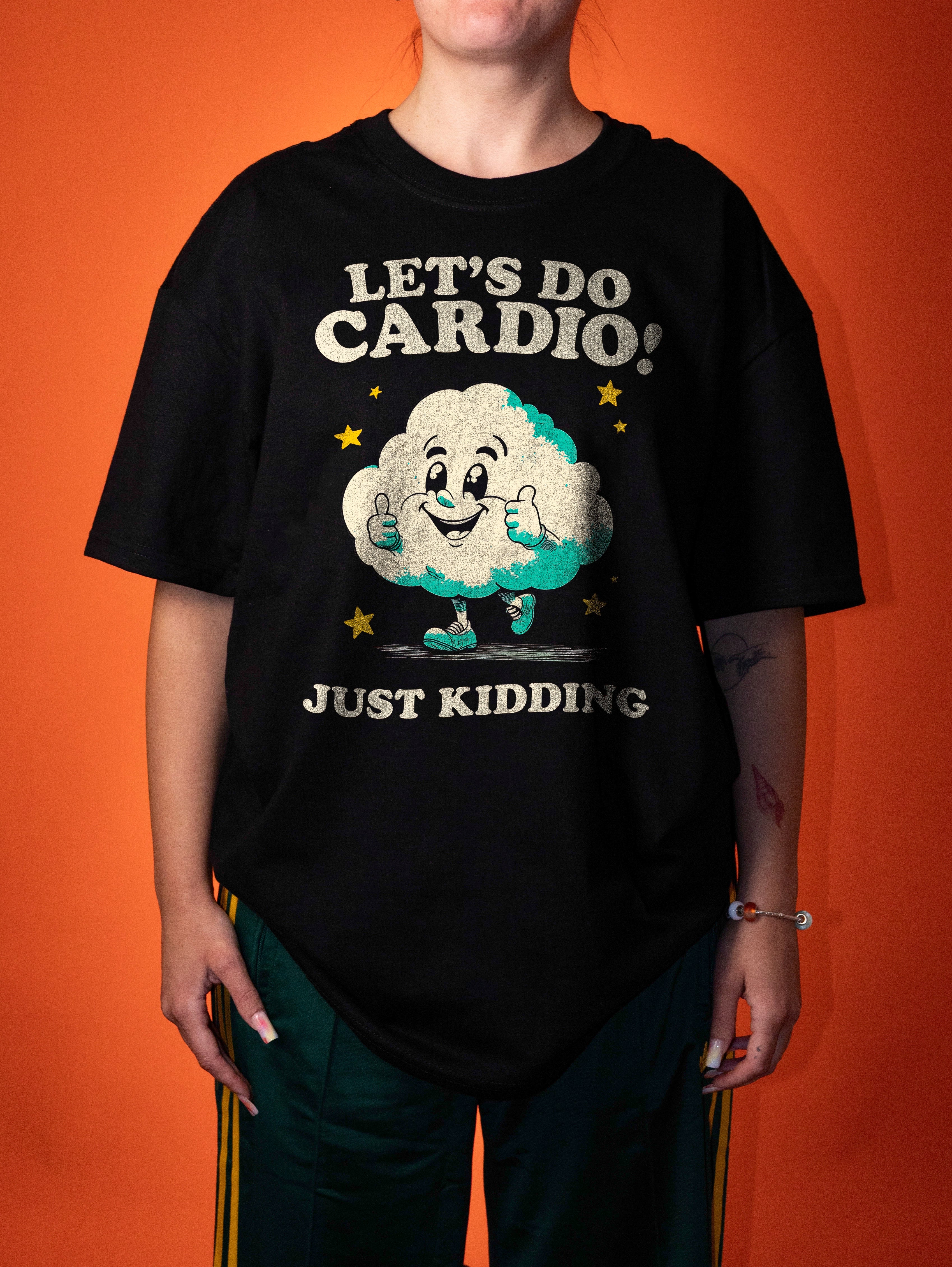 A person wearing an oversized black graphic T-shirt with a cartoon cloud and the text 'LET'S DO CARDIO! JUST KIDDING' against a solid orange background.