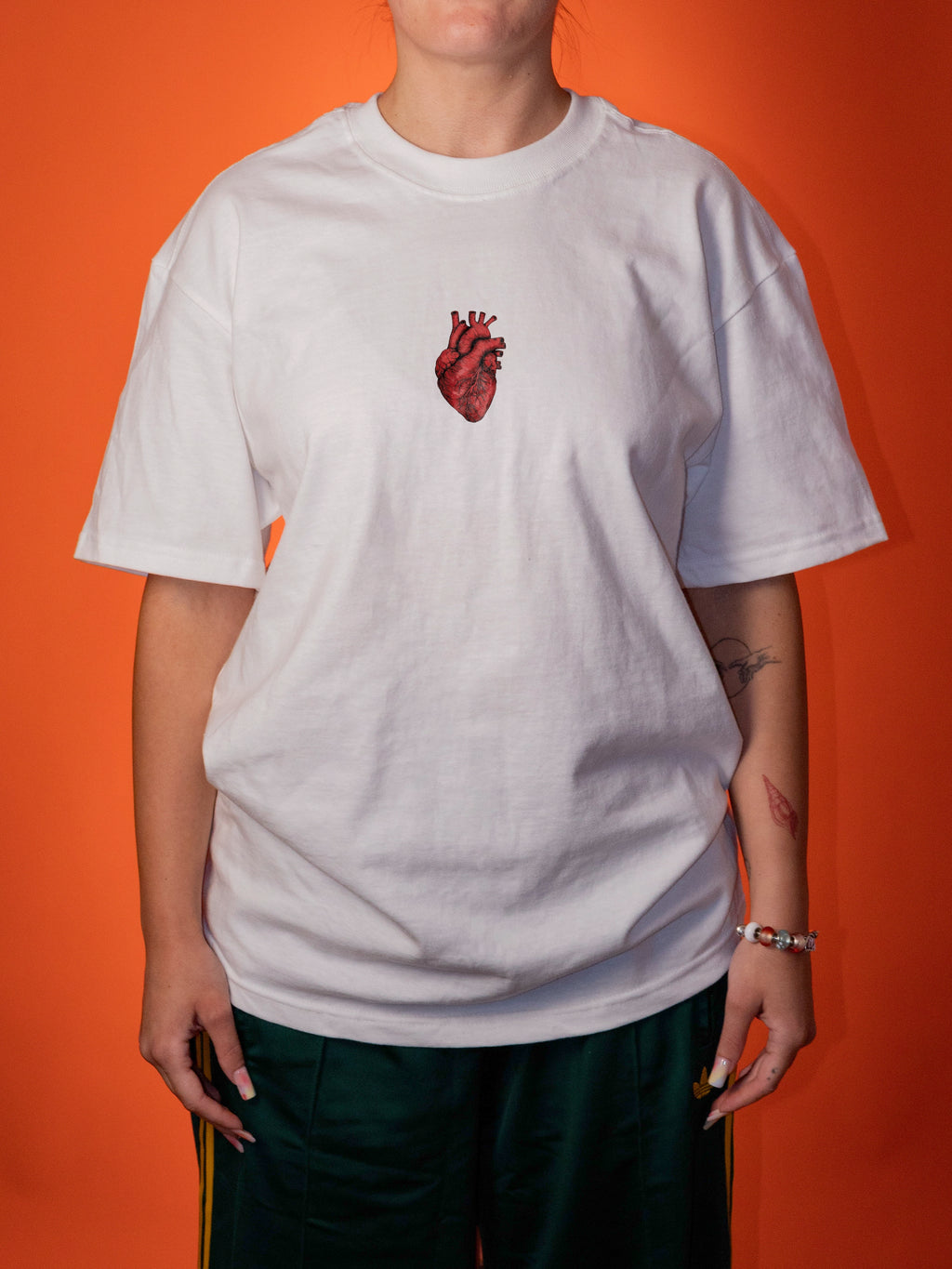 A person wearing an oversized white t-shirt with a red anatomical heart graphic on the chest, paired with dark green pants, set against a solid orange background.