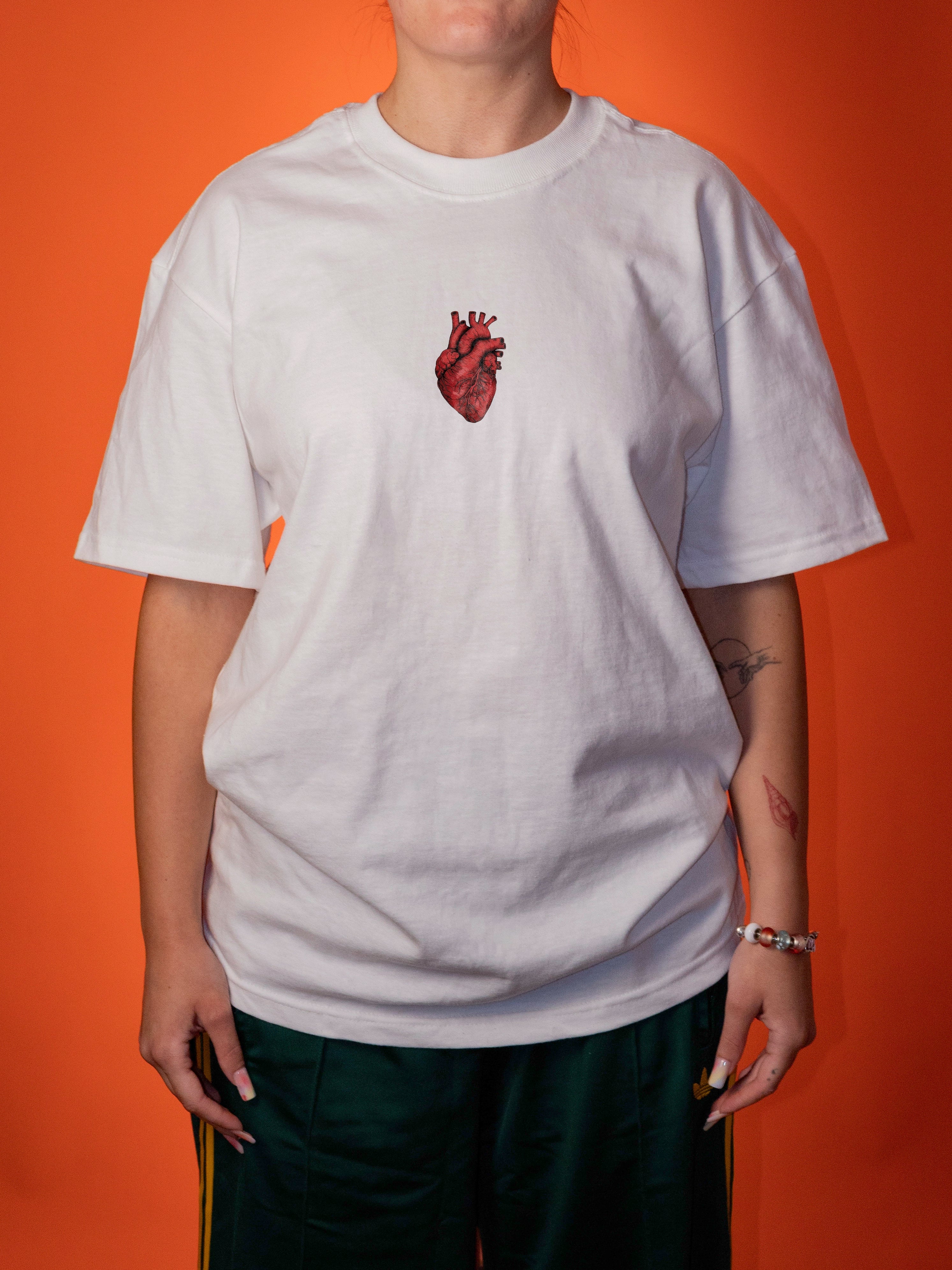 A person wearing an oversized white t-shirt with a red anatomical heart graphic on the chest, paired with dark green pants, set against a solid orange background.
