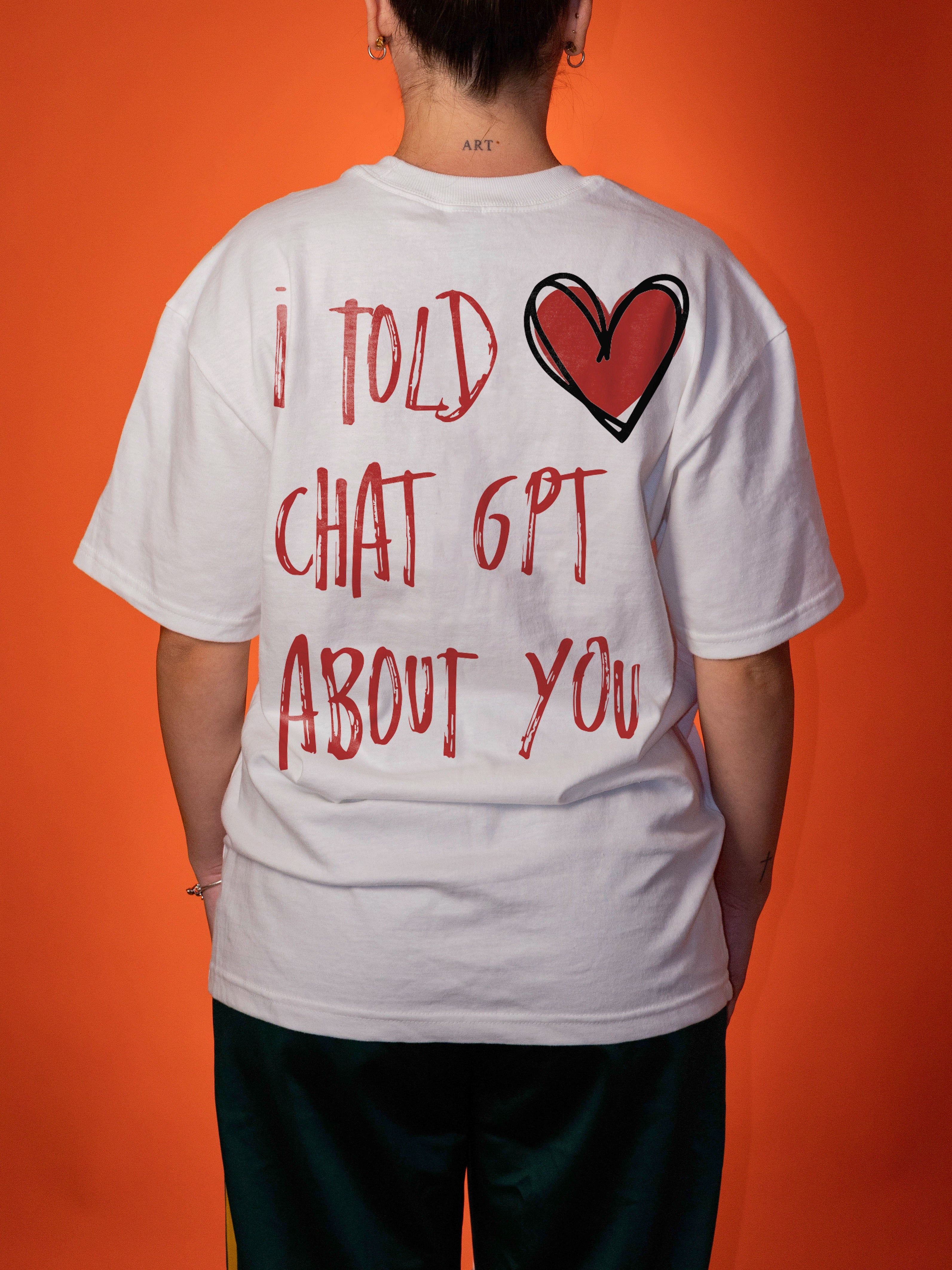 A person viewed from the back wearing a white graphic T-shirt with red hand-drawn text reading 'I TOLD CHAT GPT ABOUT YOU' and a red heart outline, against a bright orange background.