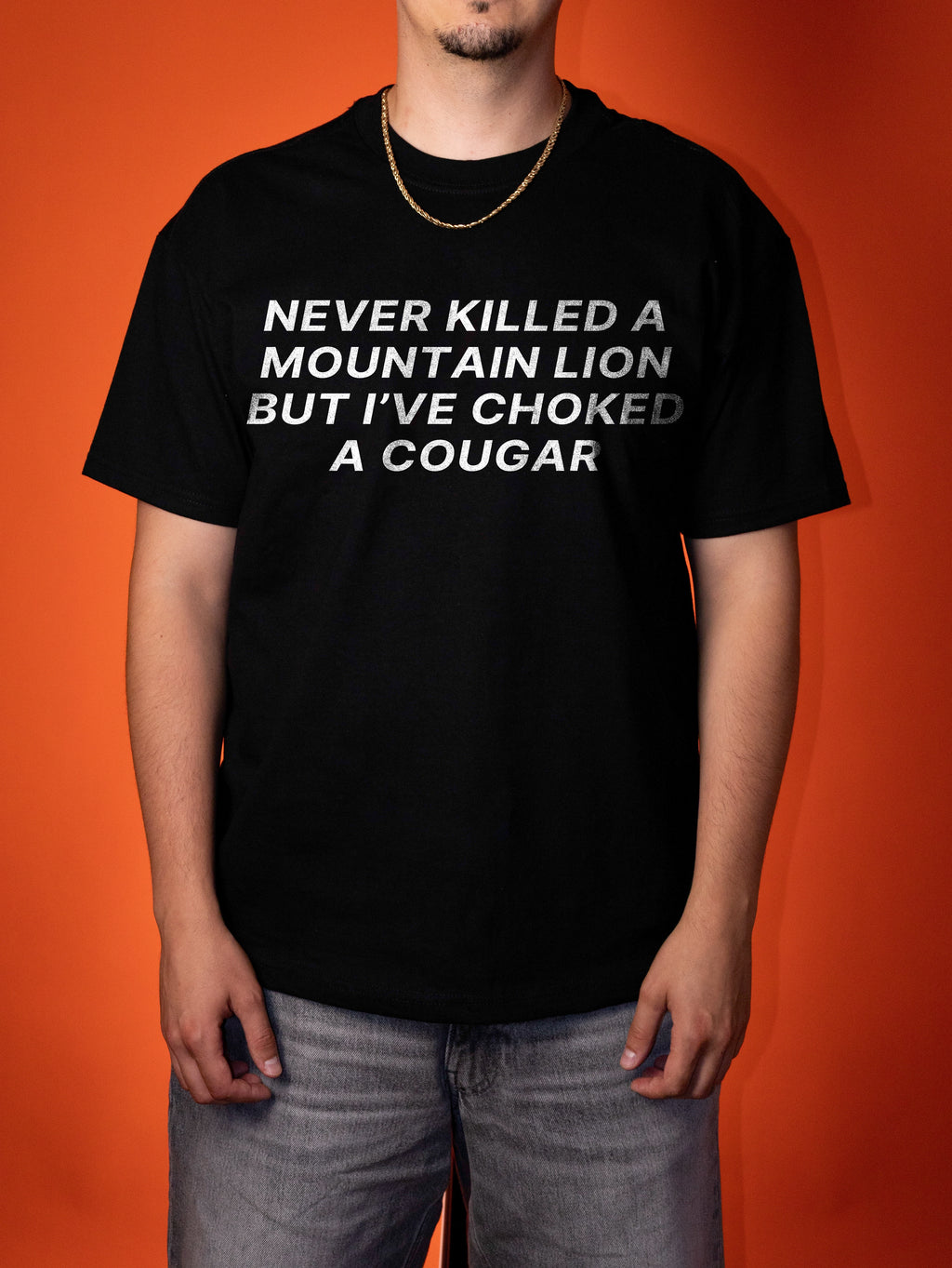 A man wearing a black graphic t-shirt with white text that reads 'NEVER KILLED A MOUNTAIN LION BUT I'VE CHOKED A COUGAR', paired with gray jeans, set against a solid orange background.