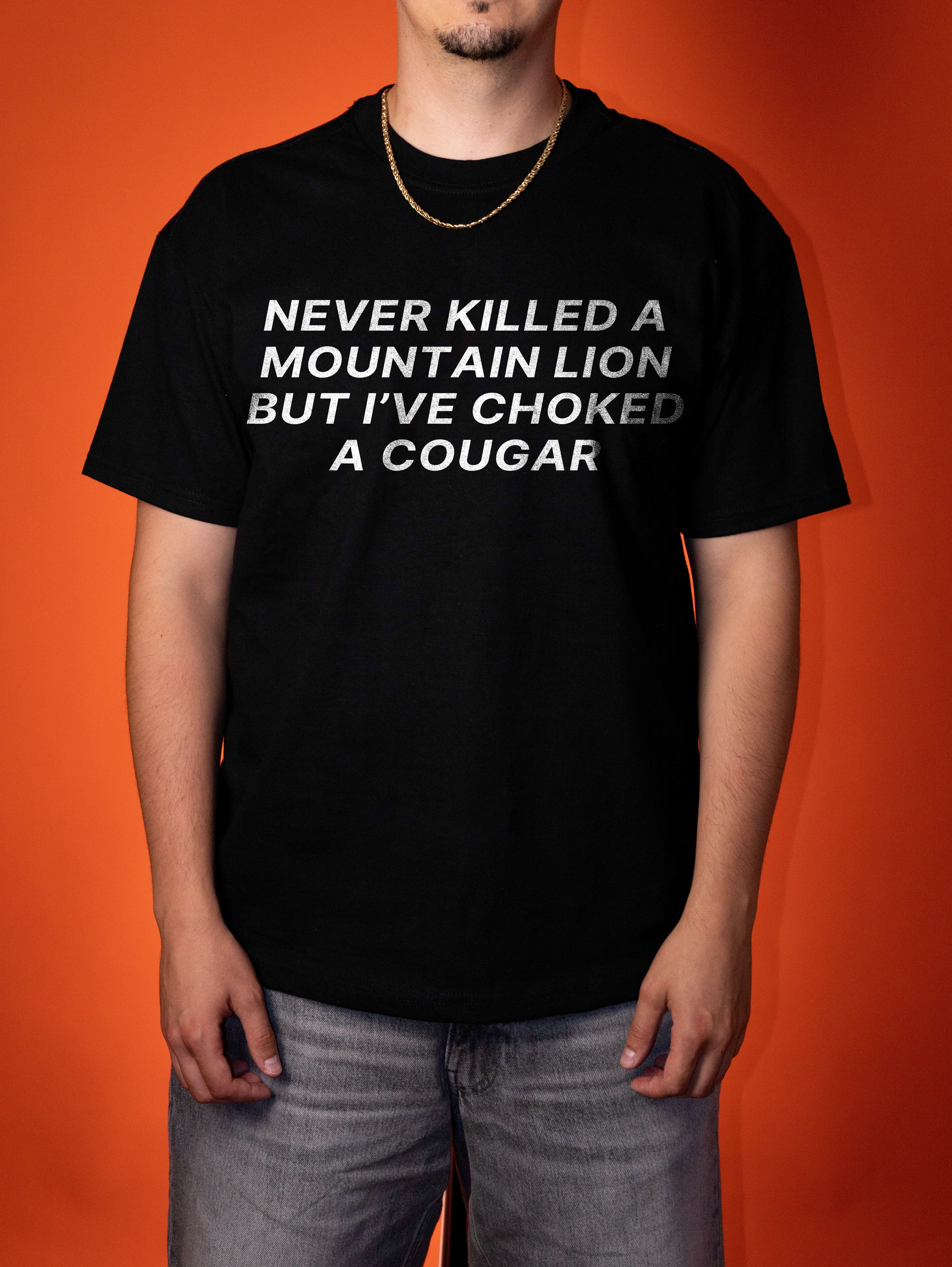A man wearing a black graphic t-shirt with white text that reads 'NEVER KILLED A MOUNTAIN LION BUT I'VE CHOKED A COUGAR', paired with gray jeans, set against a solid orange background.
