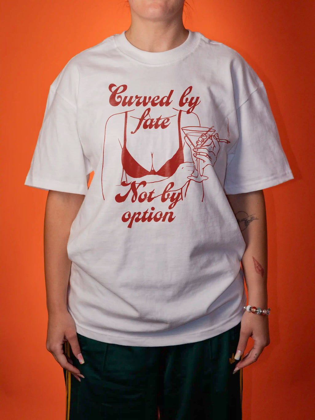 A person wearing an oversized white graphic T-shirt with red text reading 'Curved by Fate Not by option' and a red line-art illustration, posed against a solid orange background.