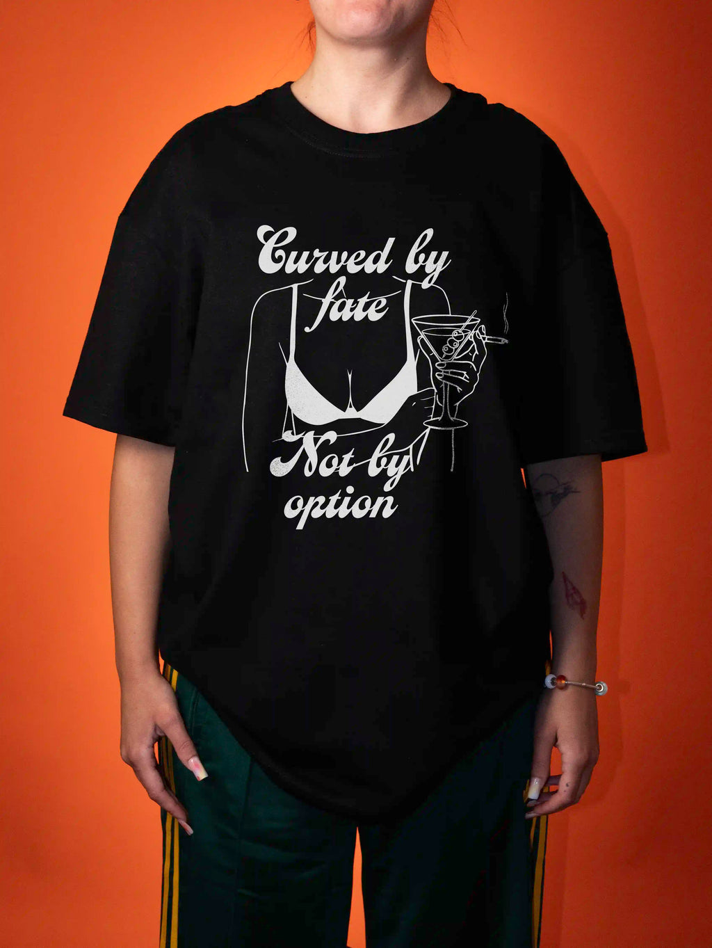 A person wearing an oversized black graphic T-shirt with white text reading 'Curved by fate Not by option' and a white illustration of a bra and martini glass, posed against a solid orange background.