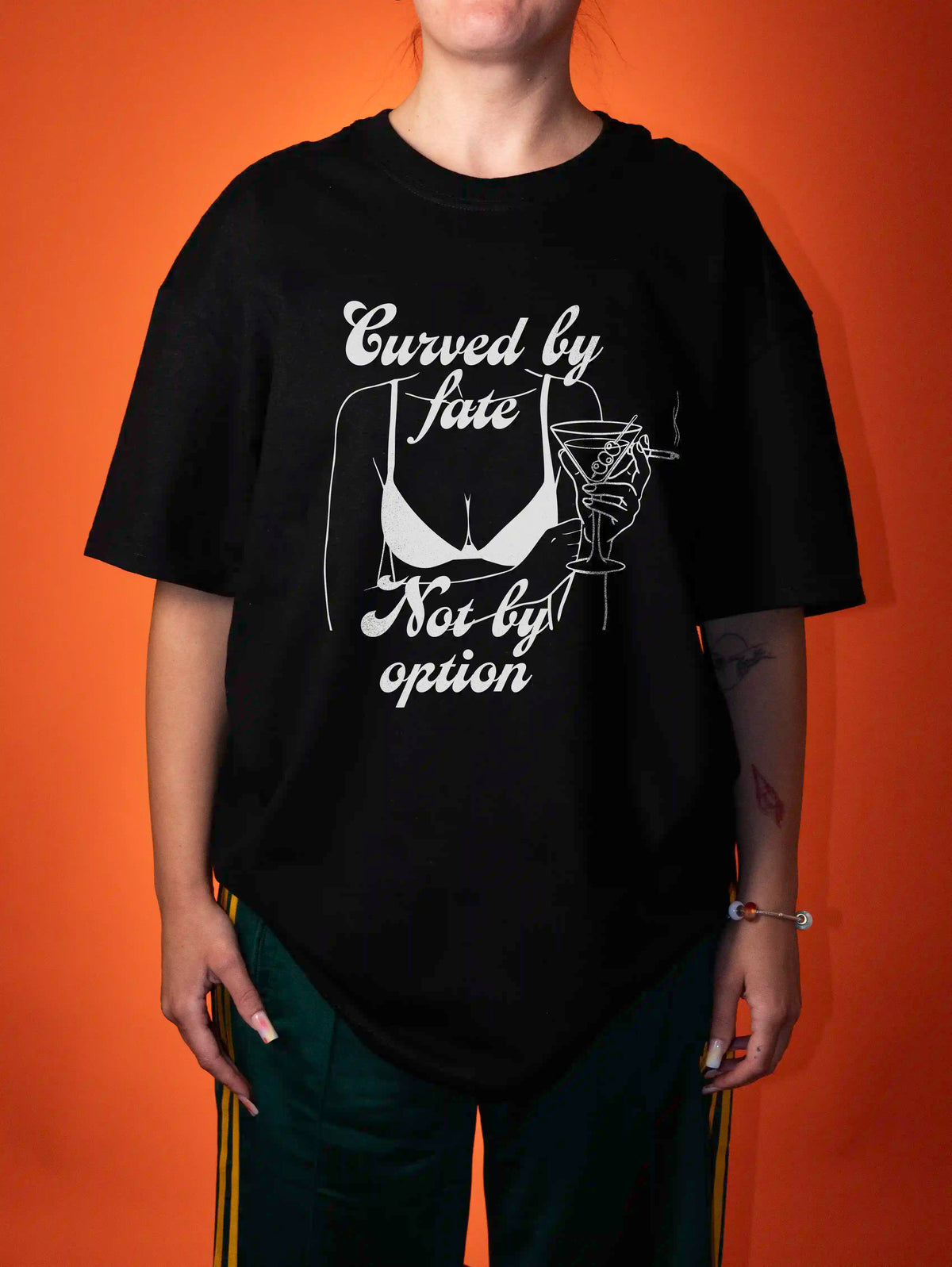 A person wearing an oversized black graphic T-shirt with white text reading 'Curved by fate Not by option' and a white illustration of a bra and martini glass, posed against a solid orange background.