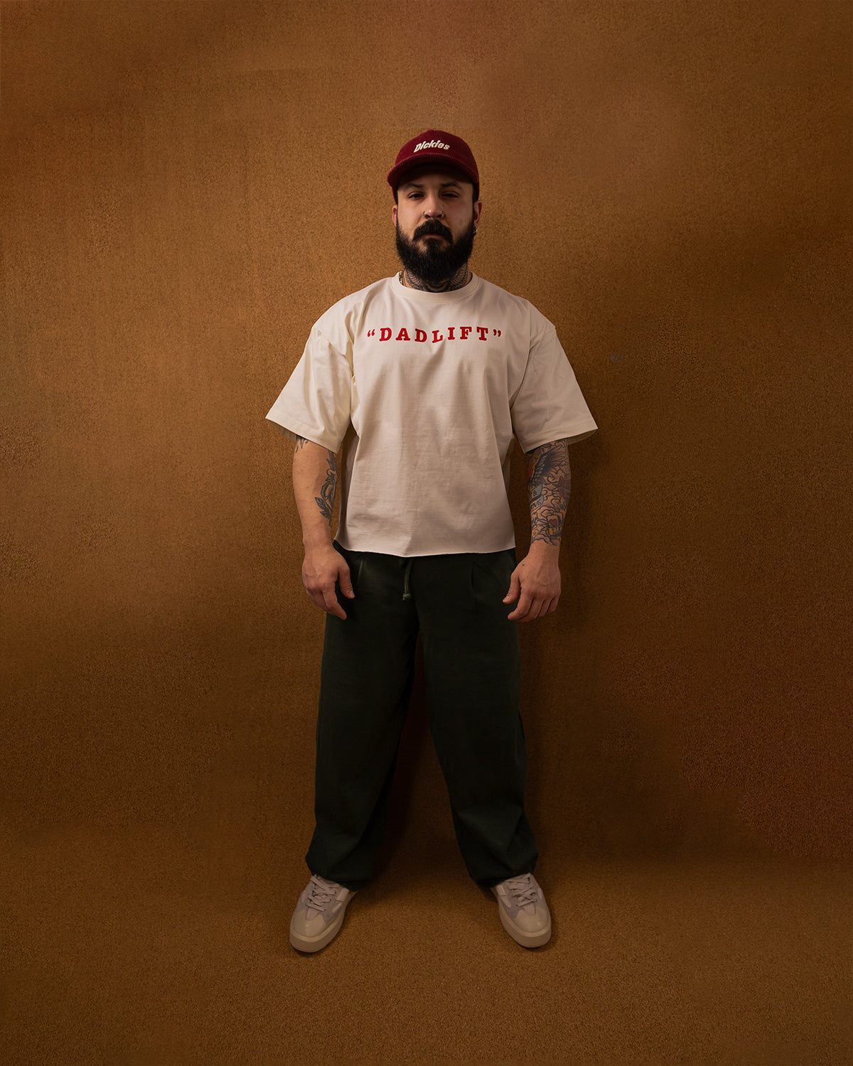 Man wearing a beige 'DADLIFT' t-shirt and dark pants against a brown background