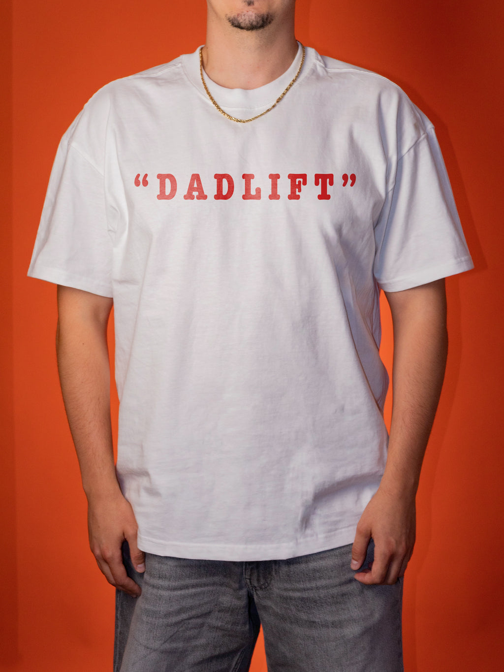 A man wearing a white short-sleeve t-shirt with 'DADLIFT' printed in red across the chest, paired with gray jeans and a gold chain necklace, set against a solid orange background.