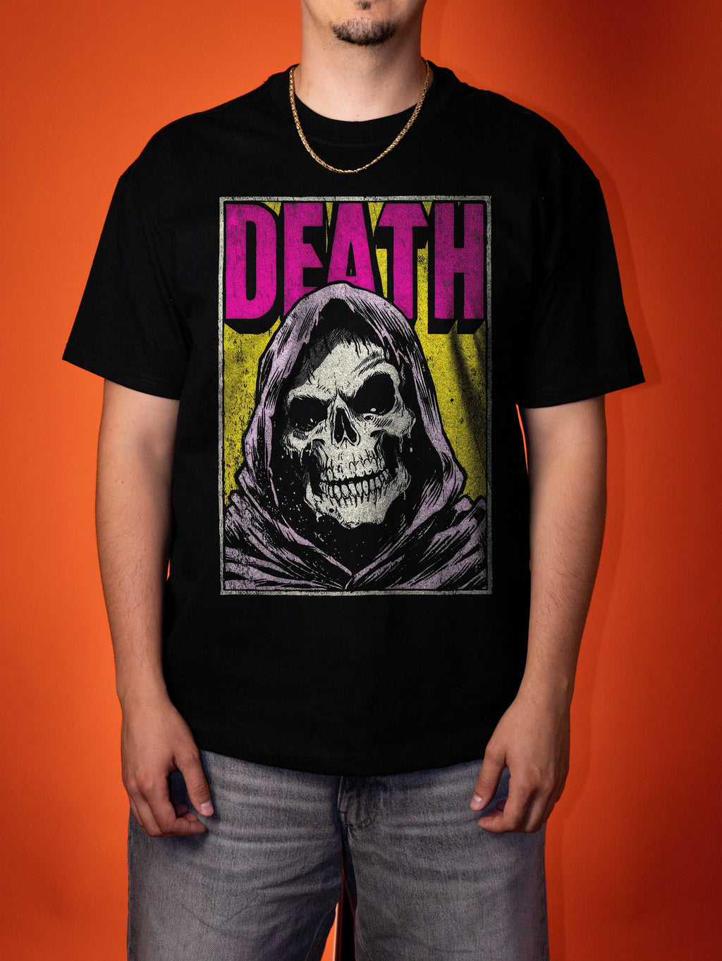 A man wearing a black graphic t-shirt featuring a grim reaper design with the word 'DEATH' in pink, set against a solid orange background.