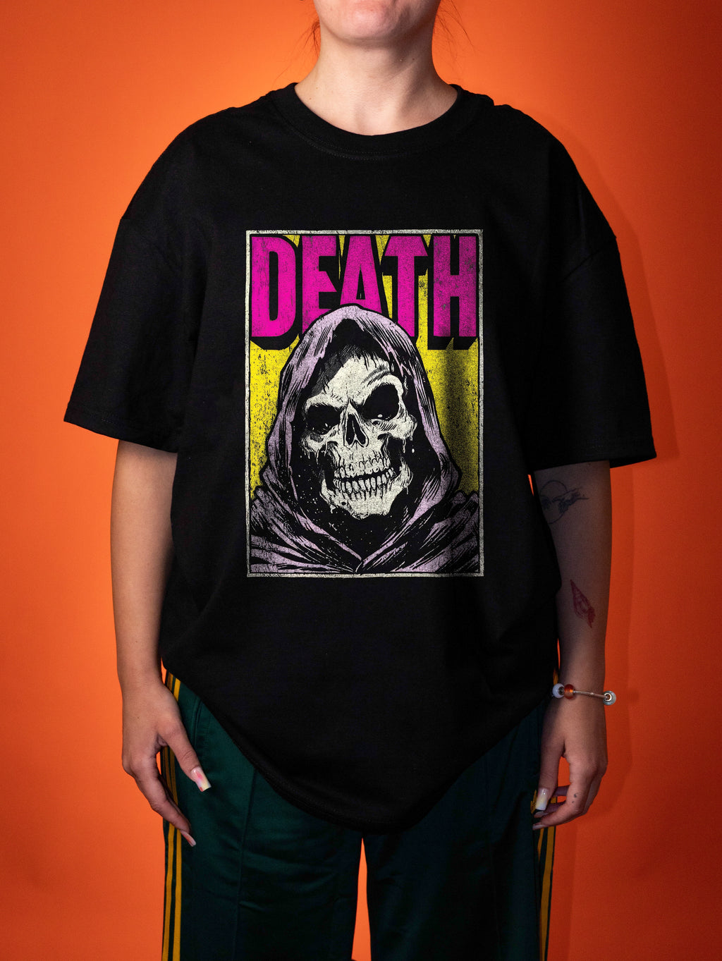 A person wearing an oversized black graphic t-shirt with a grim reaper design and the word 'DEATH' in pink, paired with dark green track pants, set against a solid orange background.
