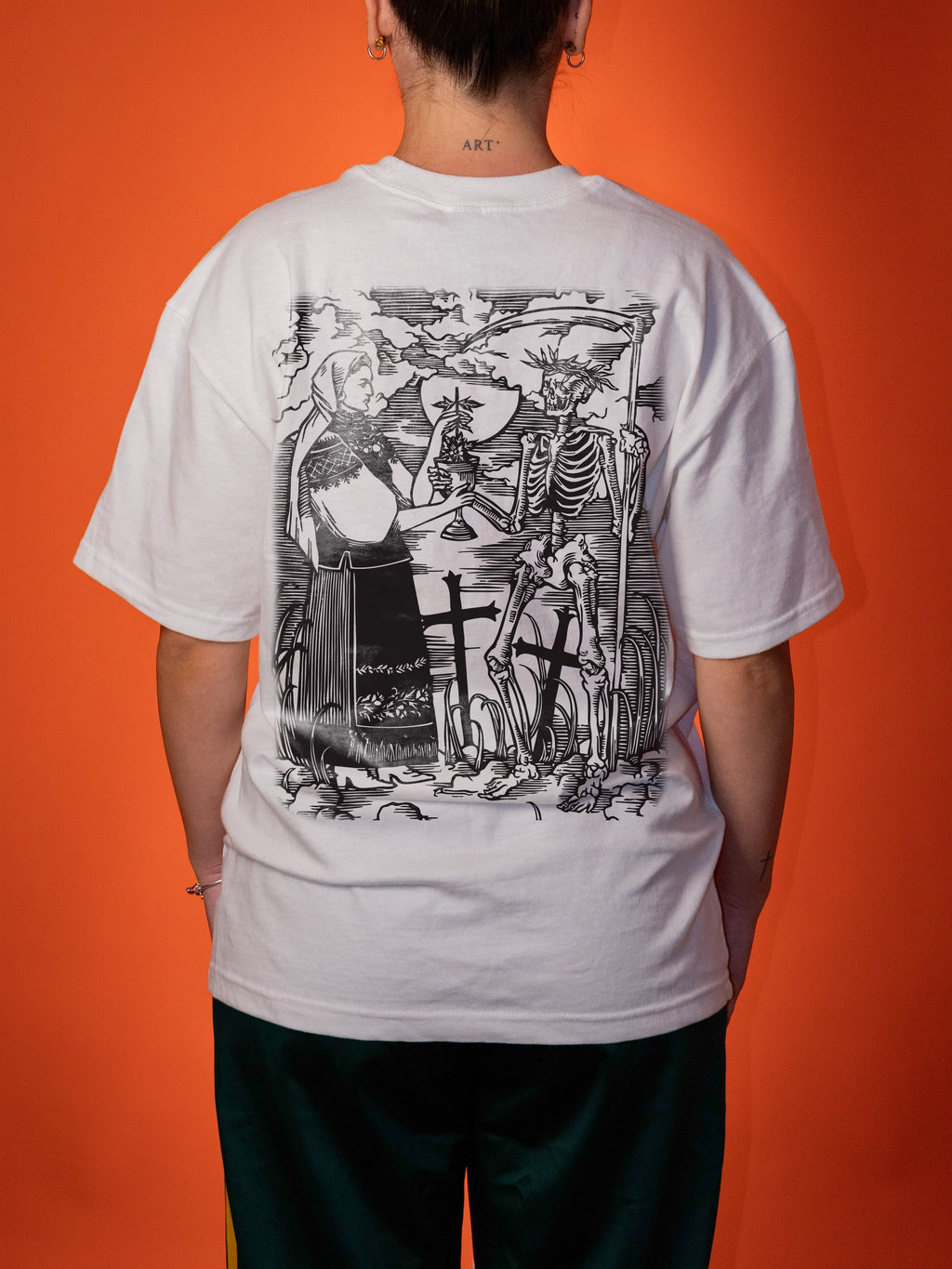 A person viewed from the back wearing a white oversized T-shirt with a large black illustrated graphic across the back, paired with dark pants, against a solid orange background.