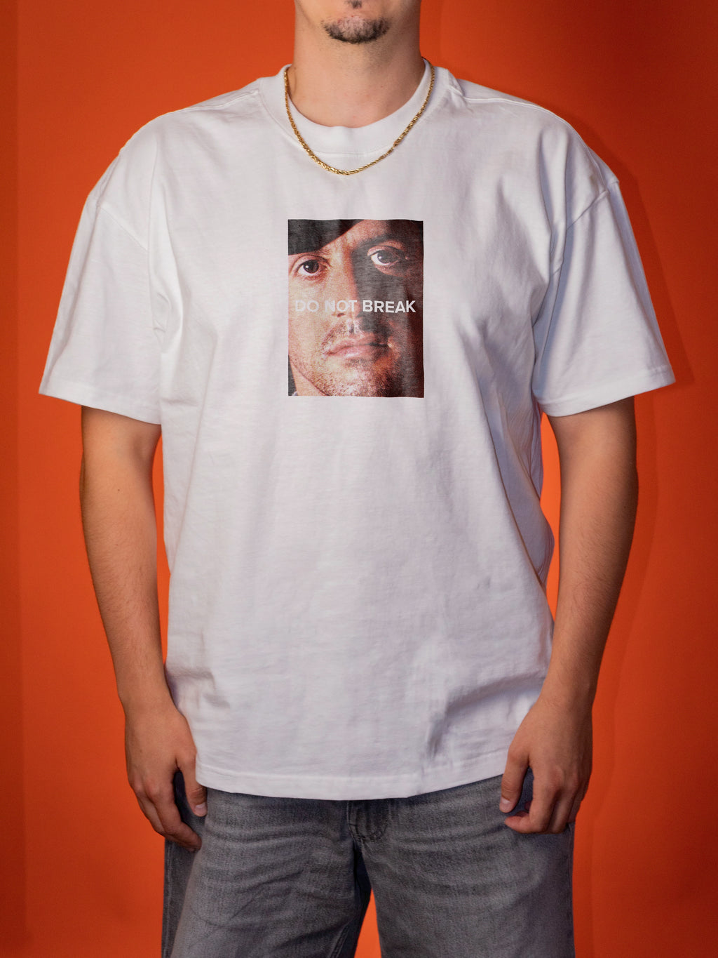 A man wearing a white graphic t-shirt with a square print of a face and the text 'DO NOT BREAK' on the chest, paired with gray jeans, set against a solid orange background.