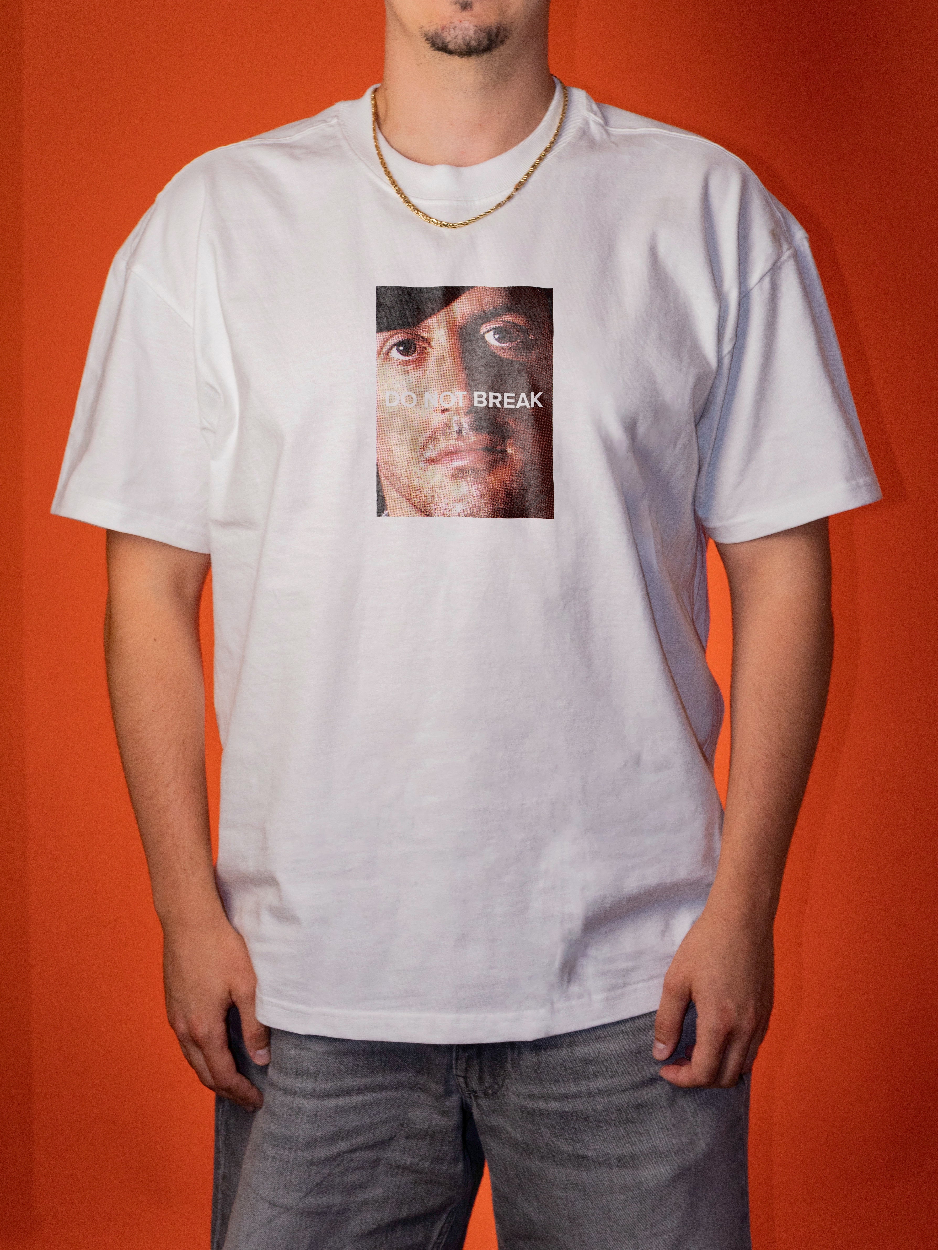 A man wearing a white graphic t-shirt with a square print of a face and the text 'DO NOT BREAK' on the chest, paired with gray jeans, set against a solid orange background.