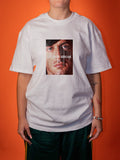 A person wearing an oversized white graphic T-shirt with a square print of a face and the text 'DO NOT BREAK' on the front, paired with dark green pants, set against a solid orange background.