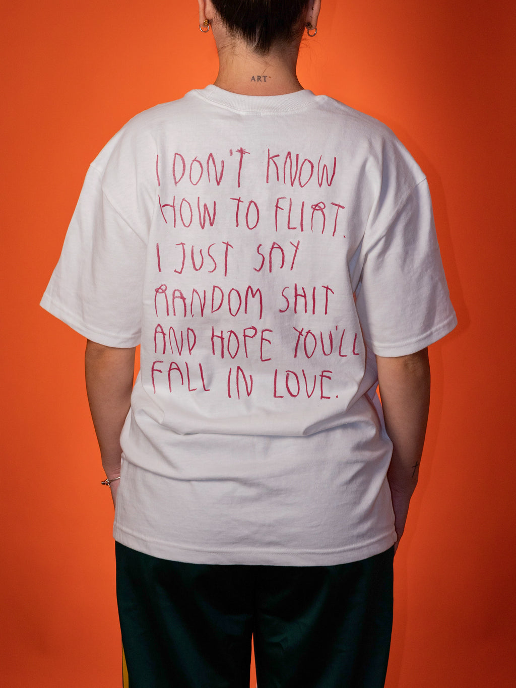 A person shown from behind wearing a white oversized T-shirt with red handwritten-style text on the back, paired with dark green pants, against a solid orange background.