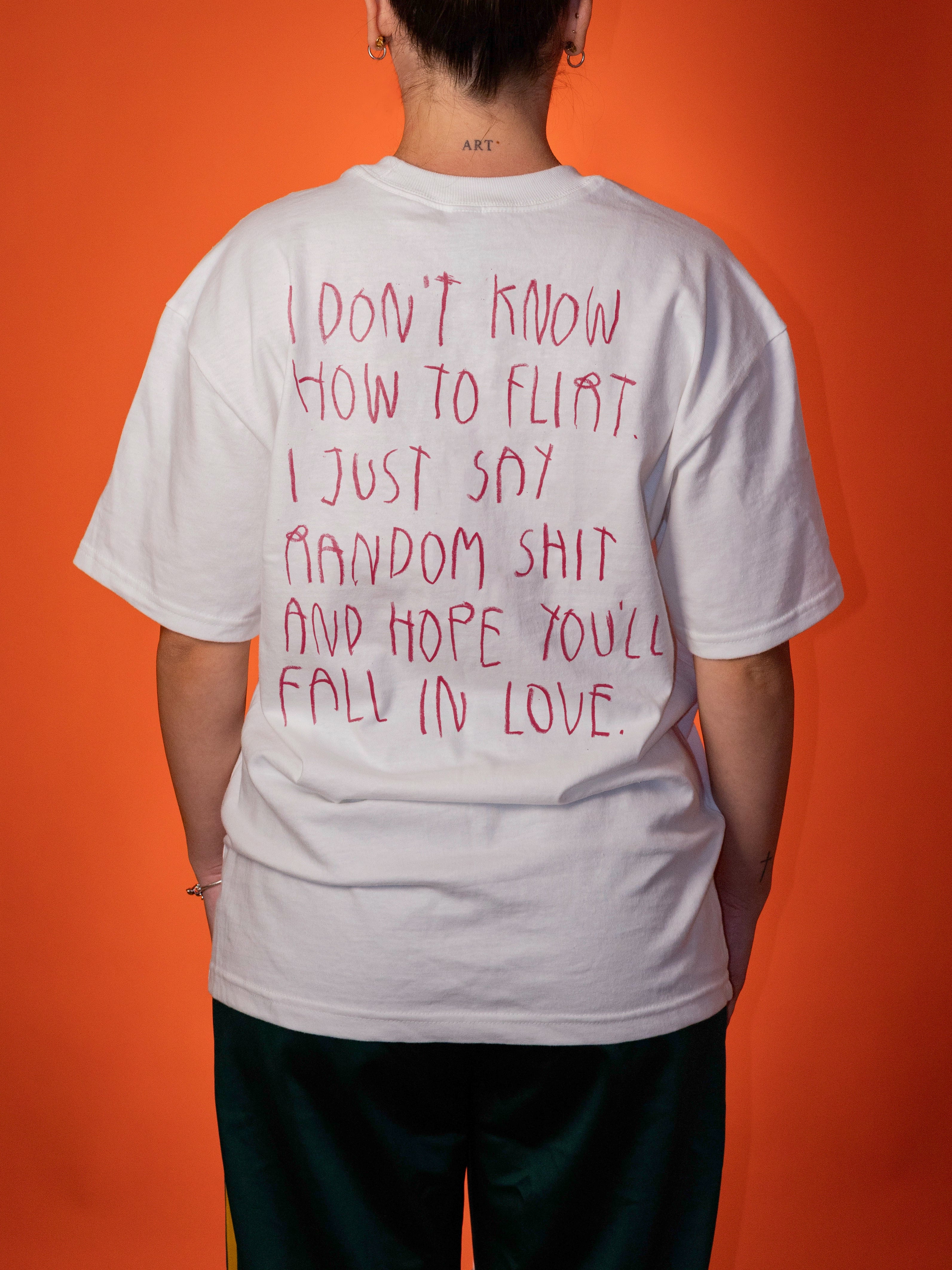 A person shown from behind wearing a white oversized T-shirt with red handwritten-style text on the back, paired with dark green pants, against a solid orange background.
