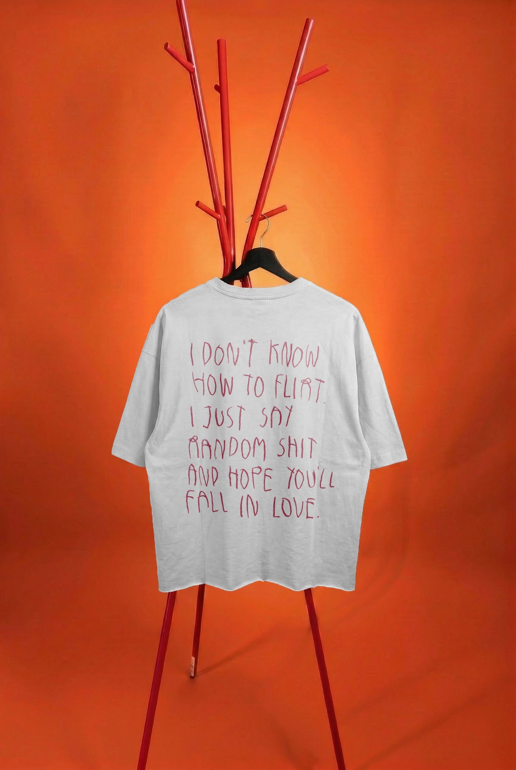A white t-shirt with pink handwritten-style text on the back, displayed on a black hanger attached to a red branch-style coat rack against a solid orange background.