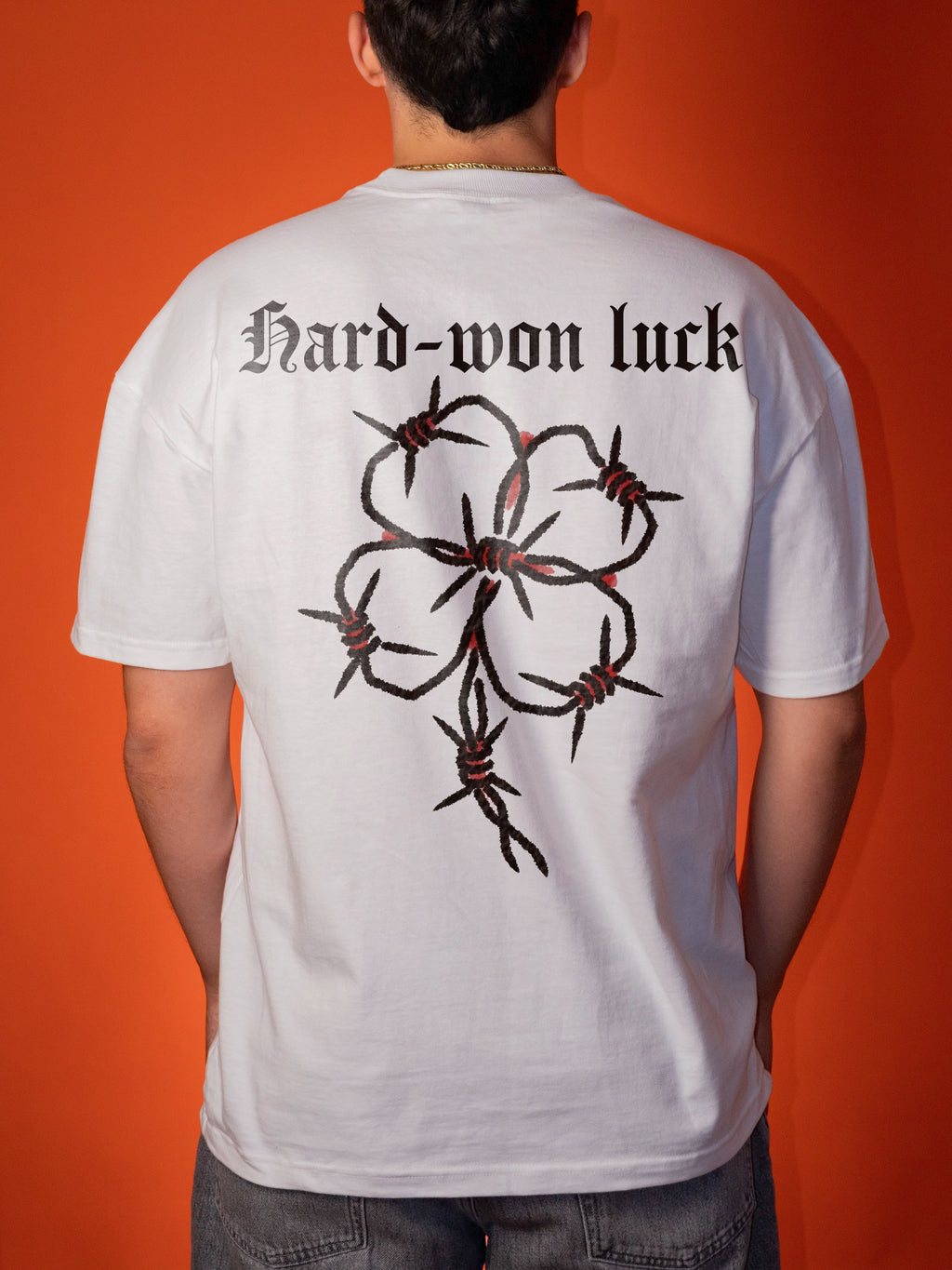 A person viewed from behind wearing a white short-sleeve T-shirt with a large black barbed-wire floral graphic and the text 'Hard-won luck' across the upper back, against an orange background.