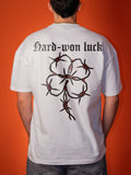 A person viewed from behind wearing a white short-sleeve T-shirt with a large black barbed-wire floral graphic and the text 'Hard-won luck' across the upper back, against an orange background.