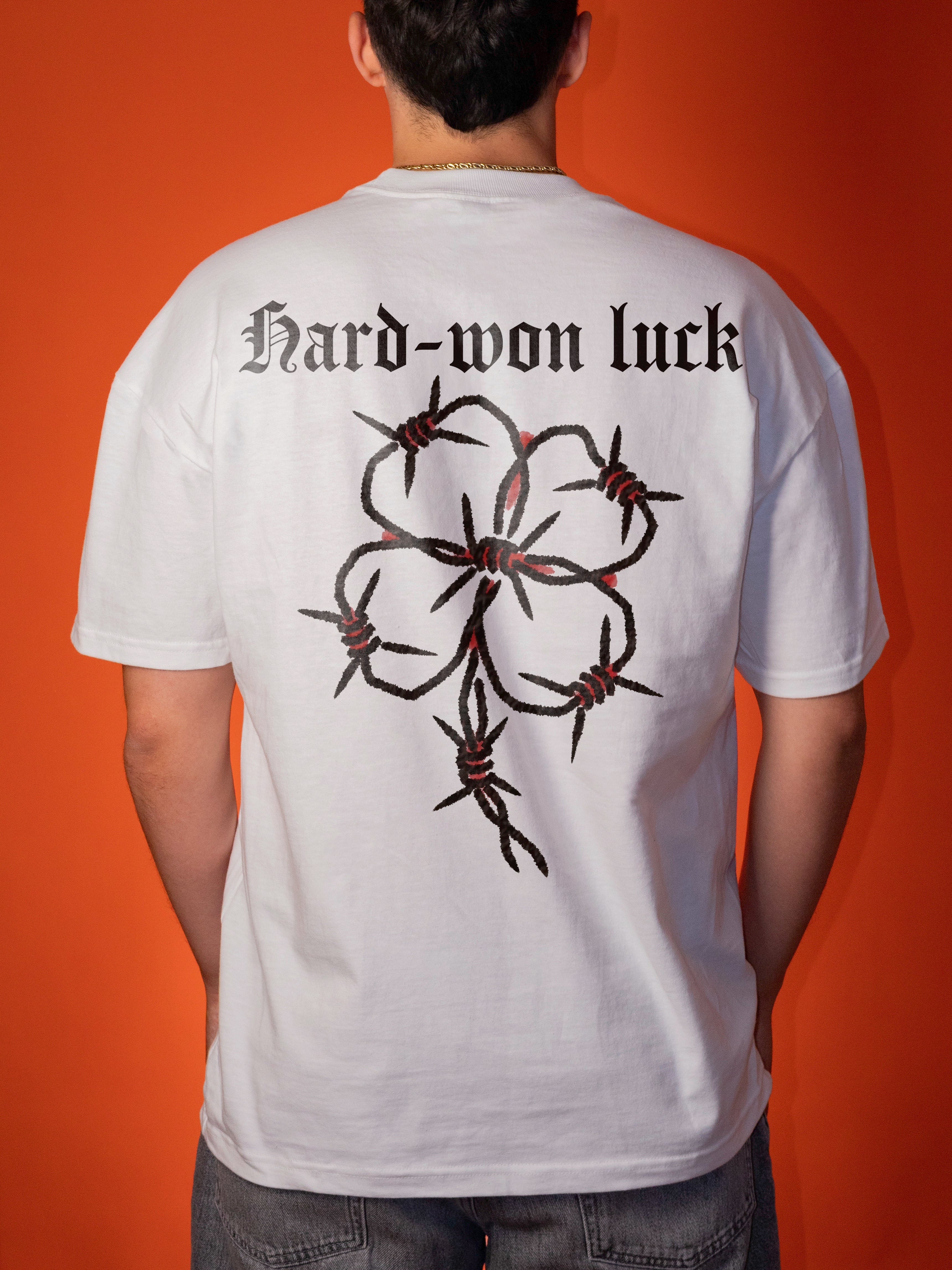 A person viewed from behind wearing a white short-sleeve T-shirt with a large black barbed-wire floral graphic and the text 'Hard-won luck' across the upper back, against an orange background.