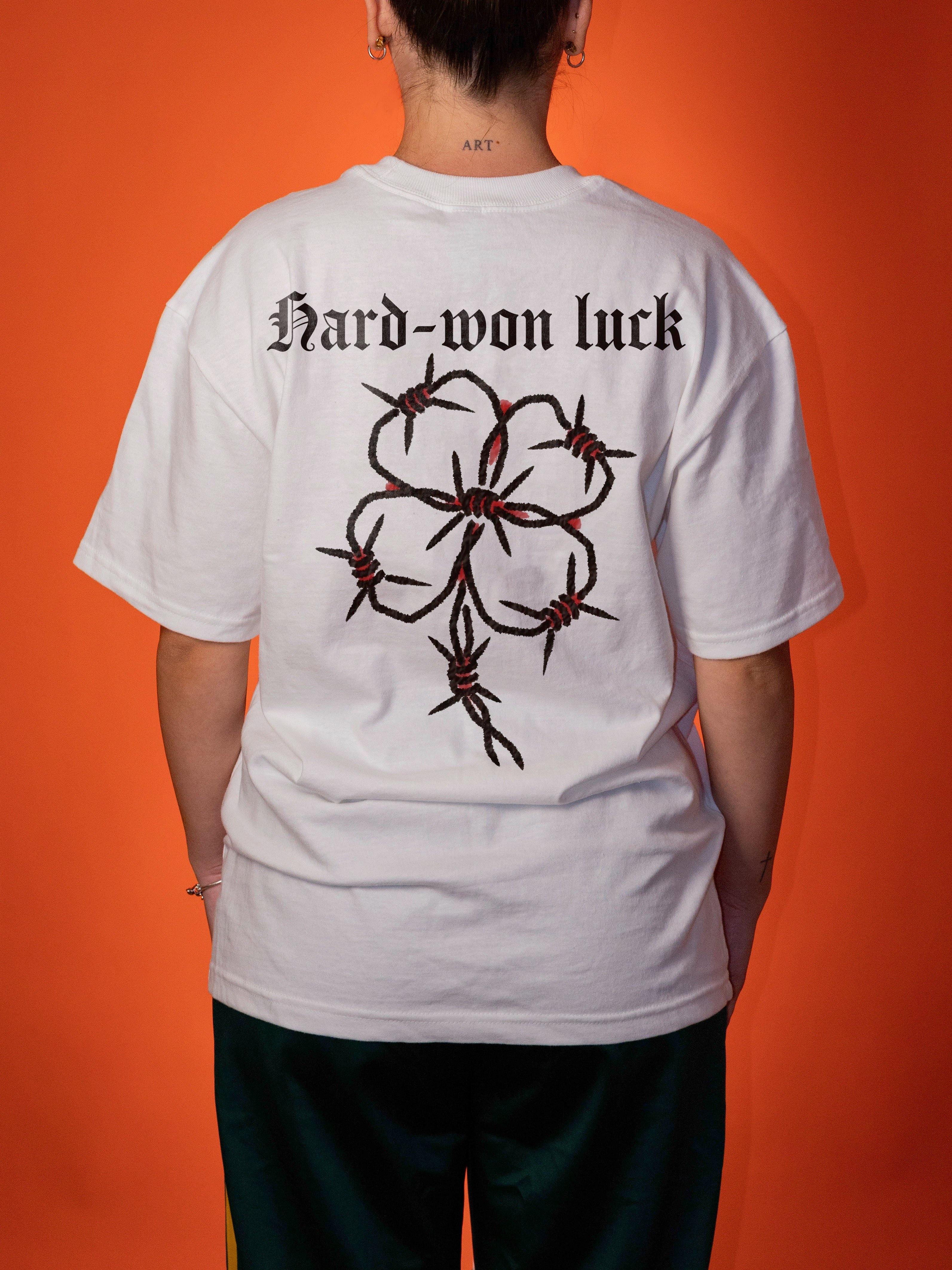 A person shown from the back wearing a white oversized T-shirt with the text 'Hard-won luck' and a large black barbed-wire flower graphic on the back, against a solid orange background.