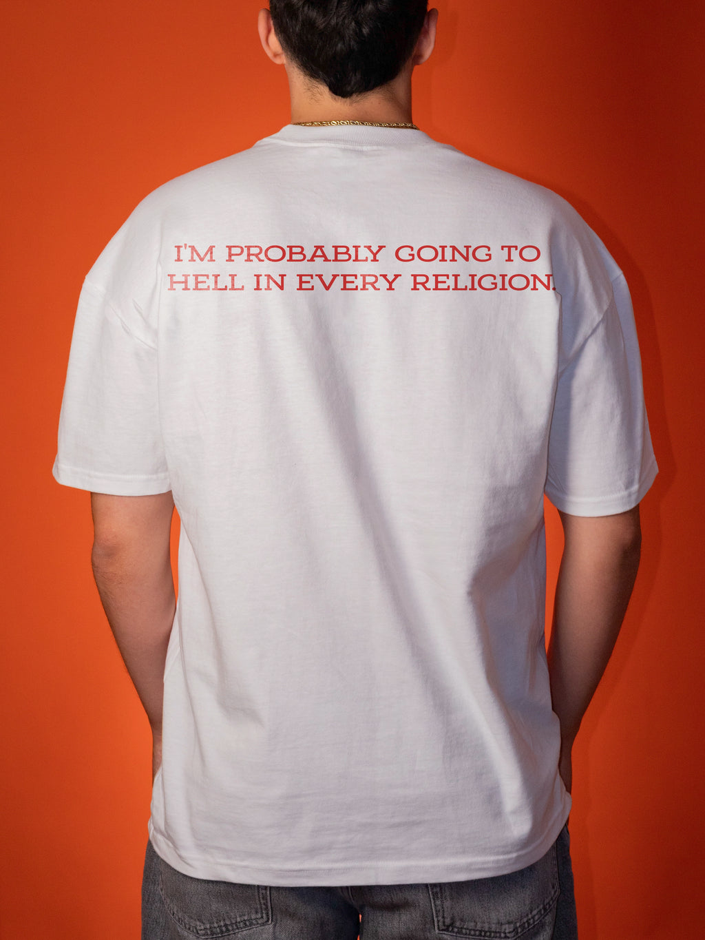 A man viewed from behind wearing a white t-shirt with red text that reads 'I'M PROBABLY GOING TO HELL IN EVERY RELIGION' against a solid orange background.
