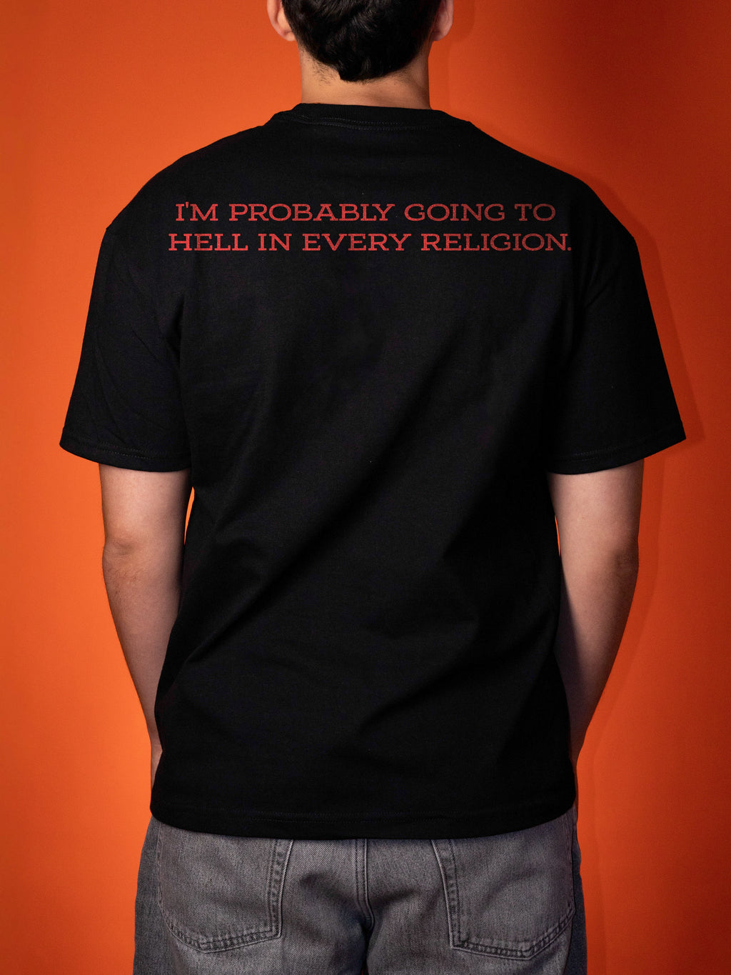 A person viewed from behind wearing a black short-sleeve T-shirt with red text on the upper back that reads, 'I'M PROBABLY GOING TO HELL IN EVERY RELIGION.' against a solid orange background.