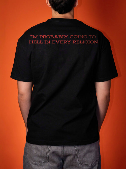 A person viewed from behind wearing a black short-sleeve T-shirt with red text on the upper back that reads, 'I'M PROBABLY GOING TO HELL IN EVERY RELIGION.' against a solid orange background.