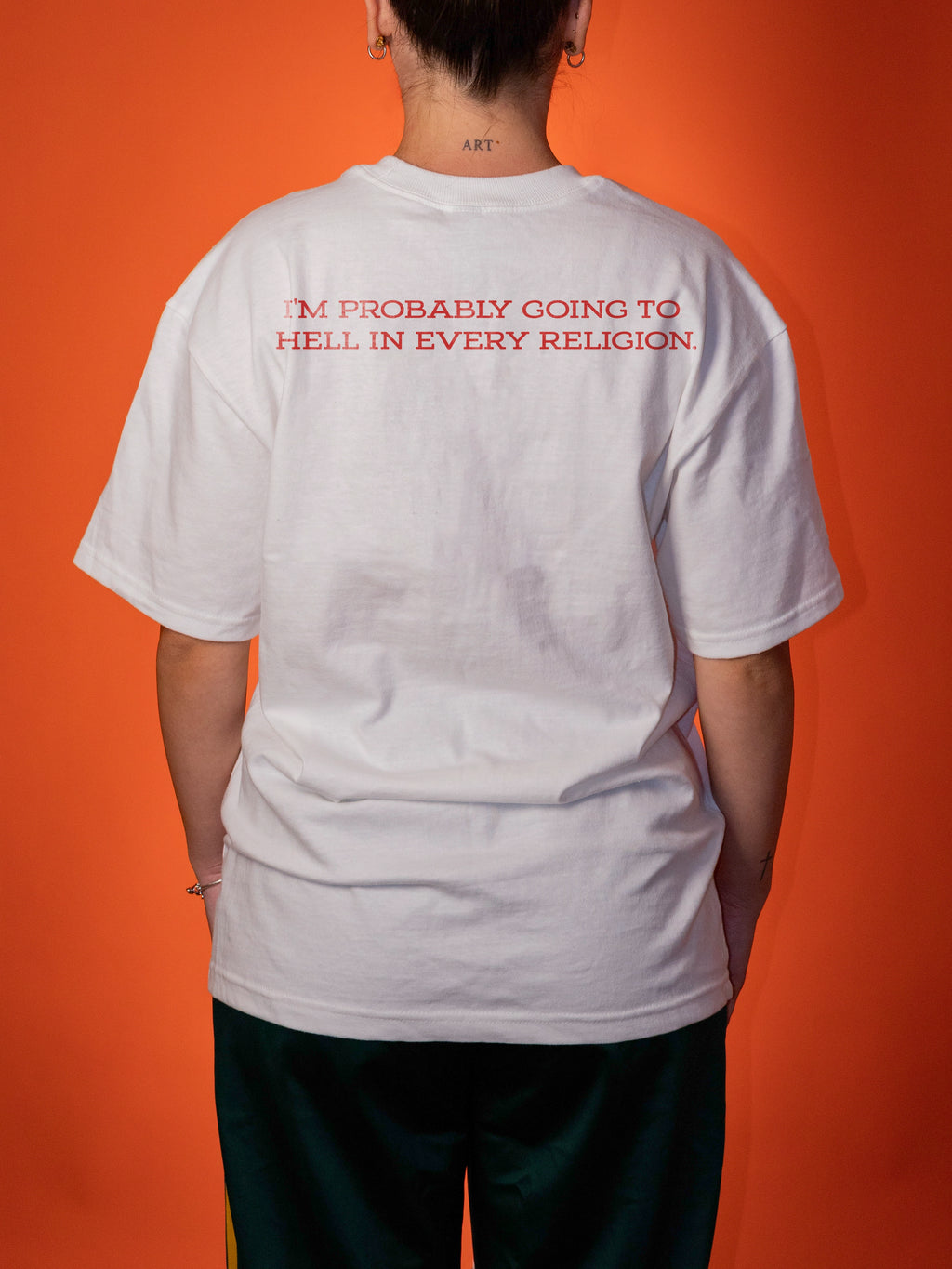 A person shown from the back wearing a white oversized T-shirt with red text across the upper back that reads, 'I'M PROBABLY GOING TO HELL IN EVERY RELIGION.' The background is a solid orange studio backdrop.