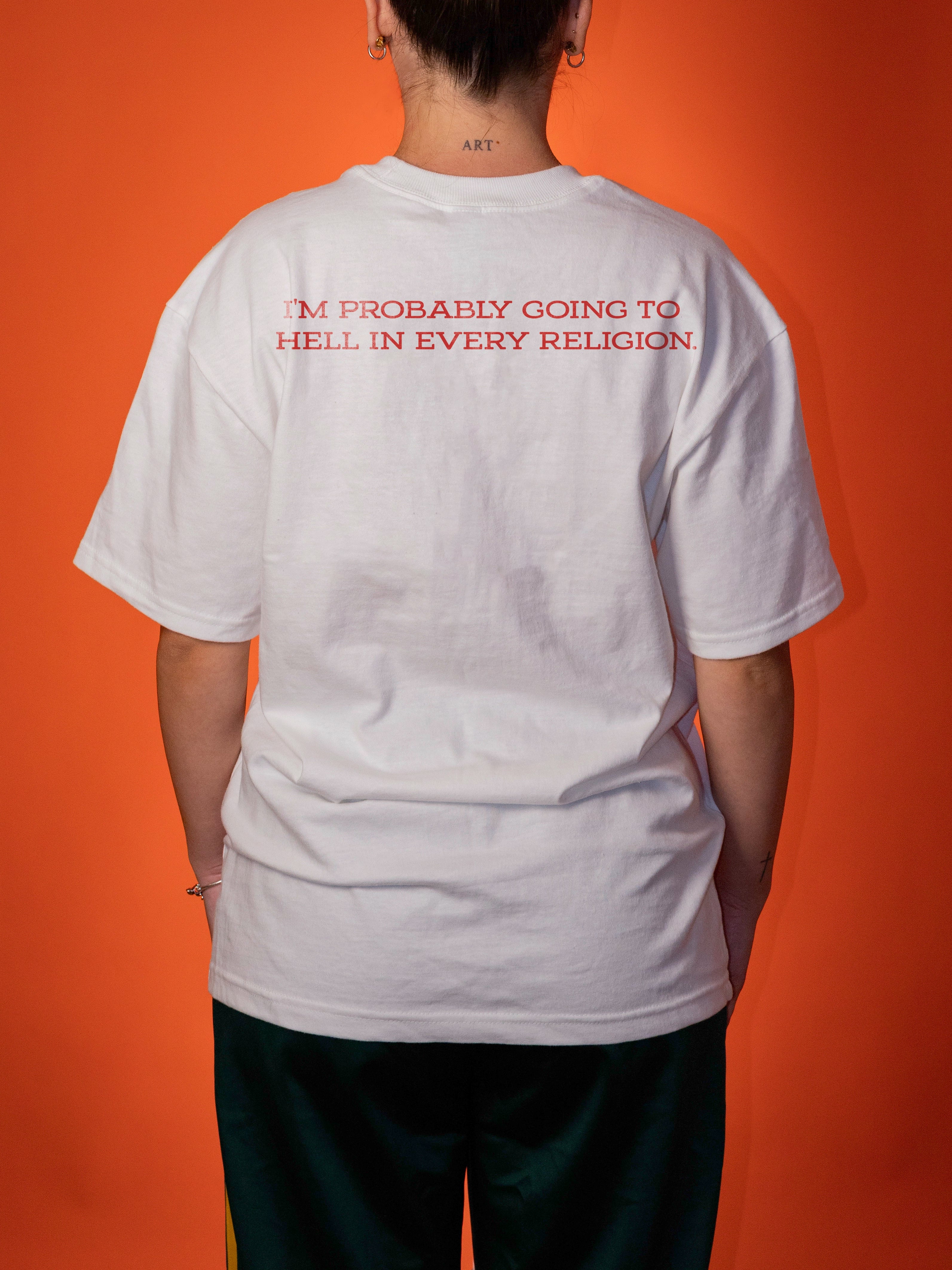 A person shown from the back wearing a white oversized T-shirt with red text across the upper back that reads, 'I'M PROBABLY GOING TO HELL IN EVERY RELIGION.' The background is a solid orange studio backdrop.