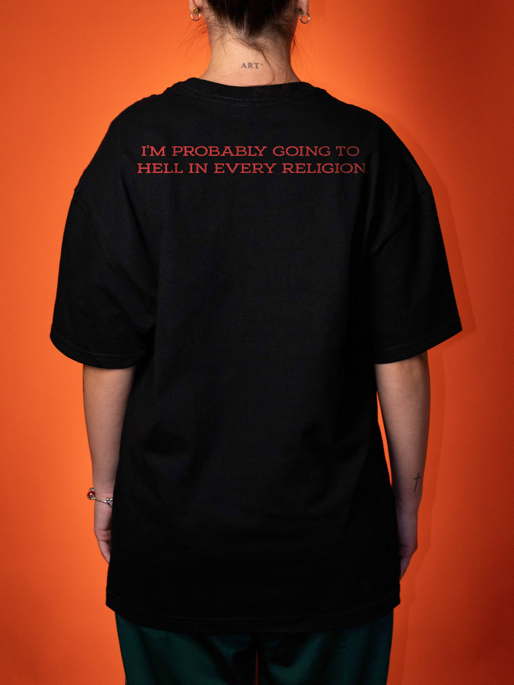 A person shown from the back wearing a black oversized T-shirt with red text across the upper back that reads, 'I'M PROBABLY GOING TO HELL IN EVERY RELIGION.' The background is a solid orange studio backdrop.