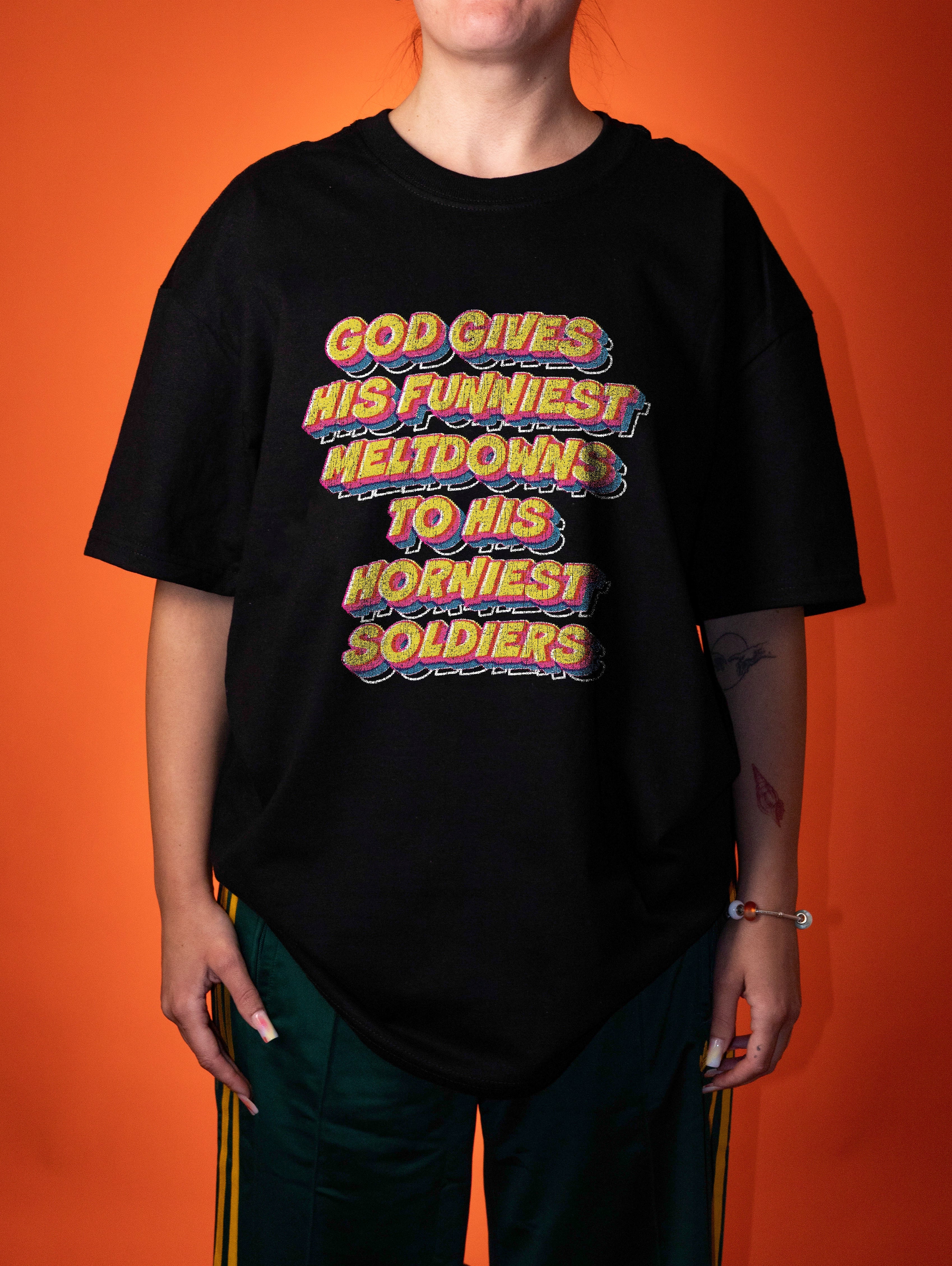 A person wearing an oversized black graphic t-shirt with a retro-style text print that reads 'GOD GIVES HIS FUNNIEST MELTDOWN TO HIS HORNIEST SOLDIERS' in yellow, orange, and purple 3D lettering, posed against a solid orange background.