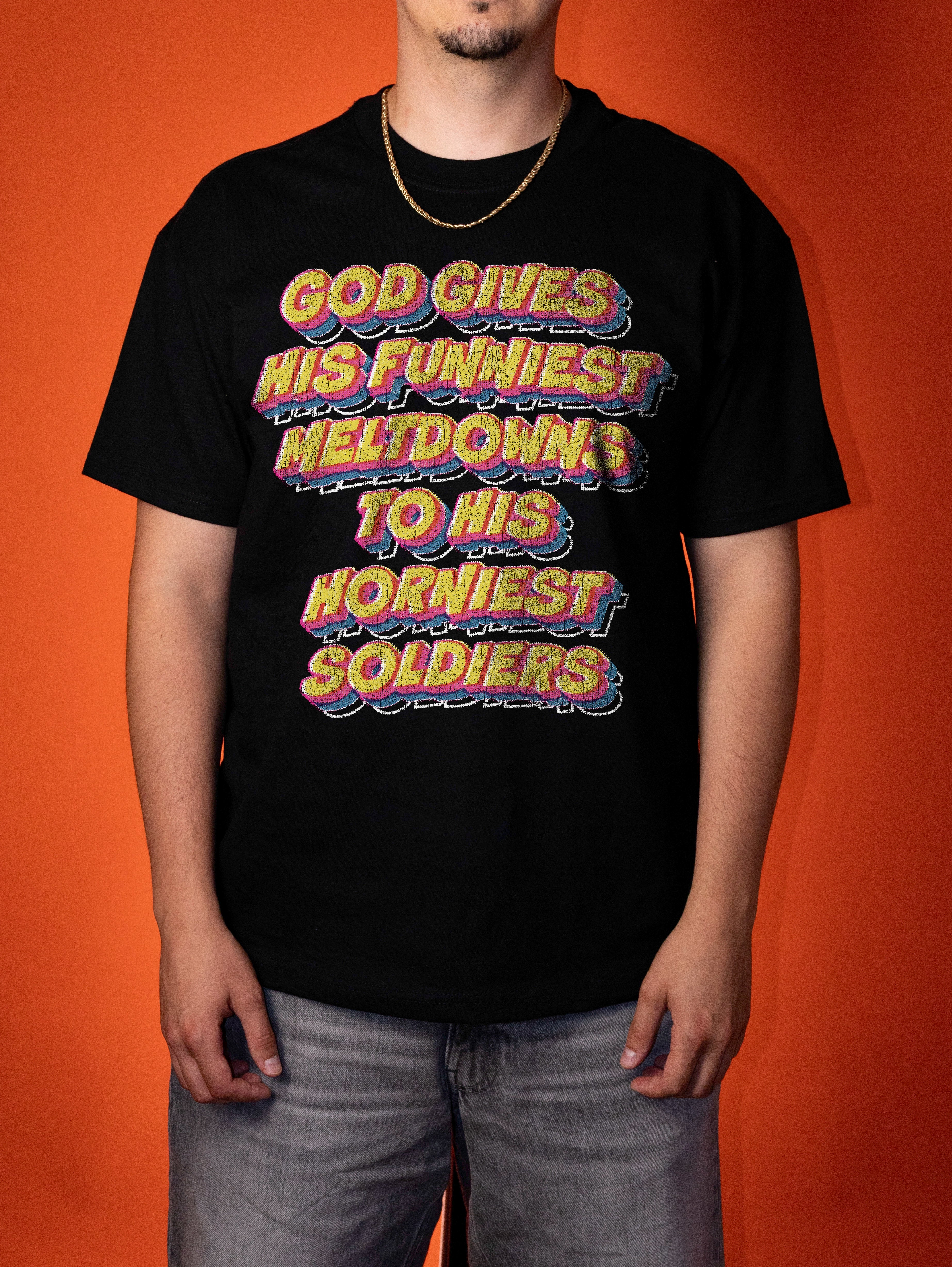 A man wearing a black graphic t-shirt with a colorful retro-style text design that reads 'GOD GIVES HIS FUNNIEST MELTDOWN TO HIS HORNINIEST SOLDIERS', set against a solid orange background.