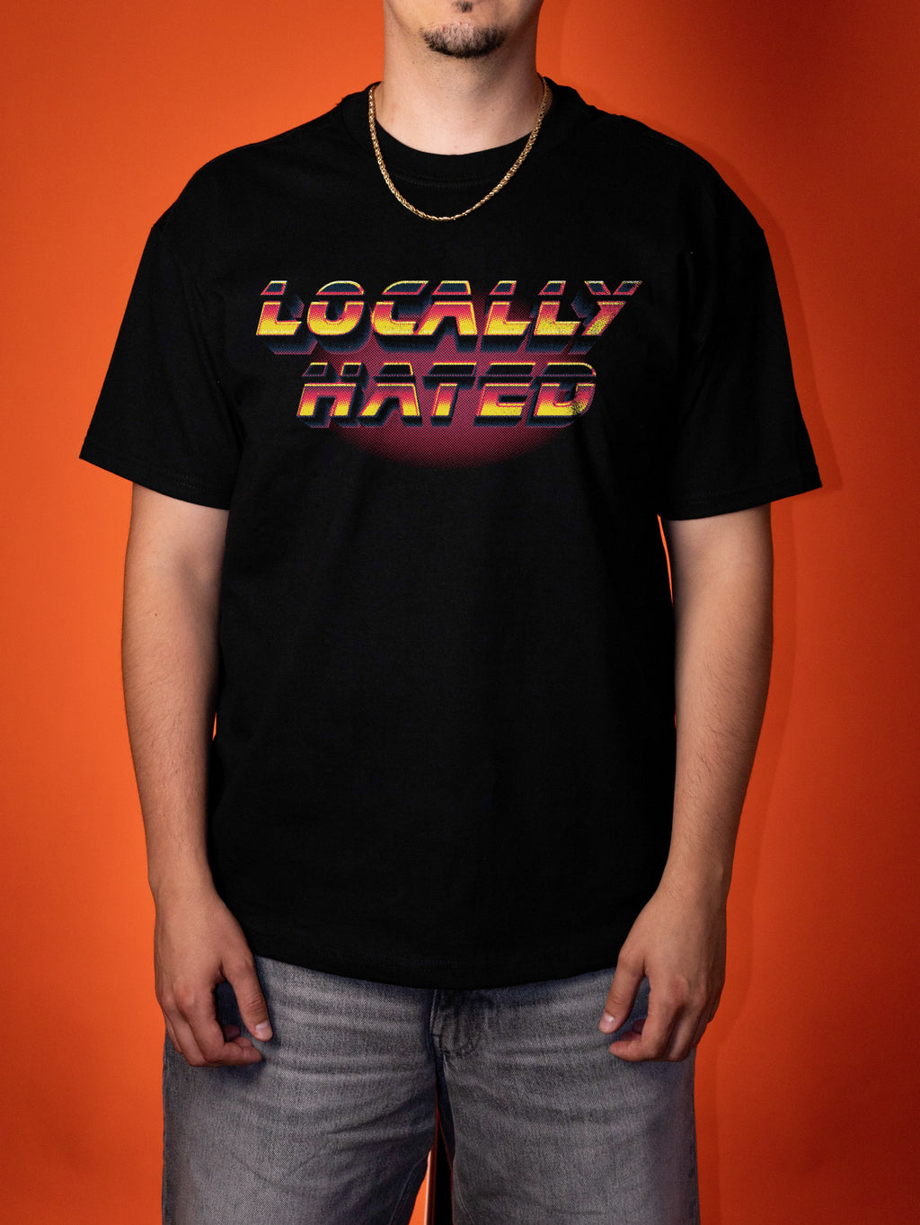 A man wearing a black t-shirt with a retro-style 'LOCALLY HATED' graphic in yellow, orange, and red gradient text, standing against a solid orange background.