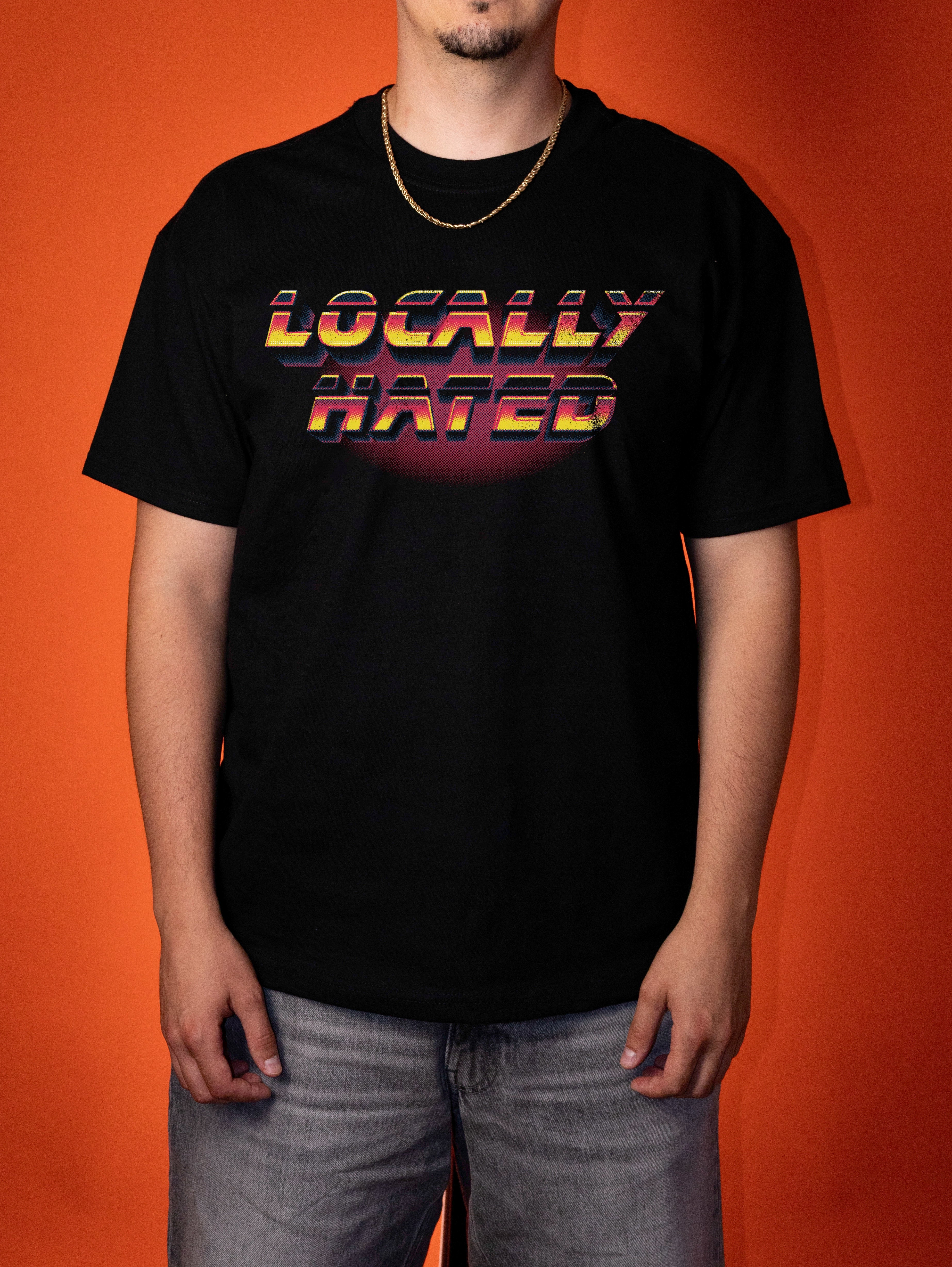 A man wearing a black t-shirt with a retro-style 'LOCALLY HATED' graphic in yellow, orange, and red gradient text, standing against a solid orange background.