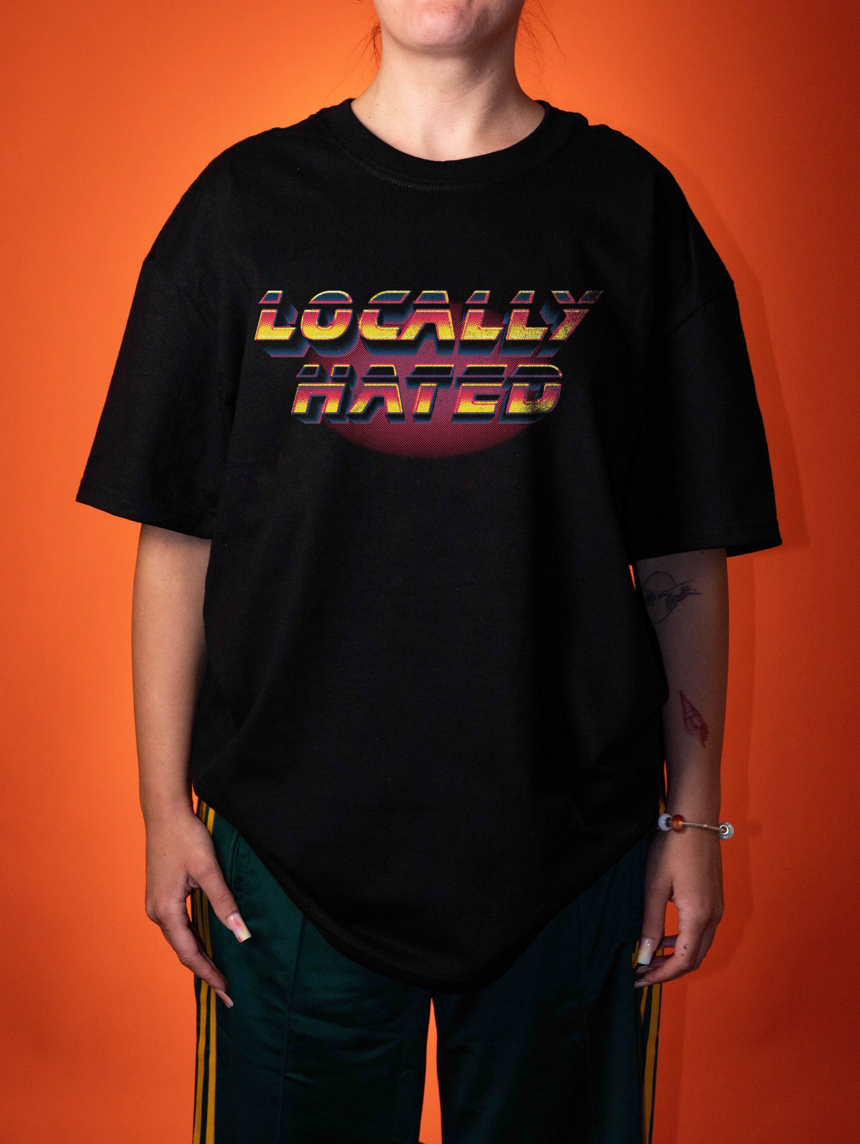 A person wearing an oversized black graphic T-shirt with the words 'LOCALLY HATED' in a retro rainbow gradient, posed against a solid orange background.